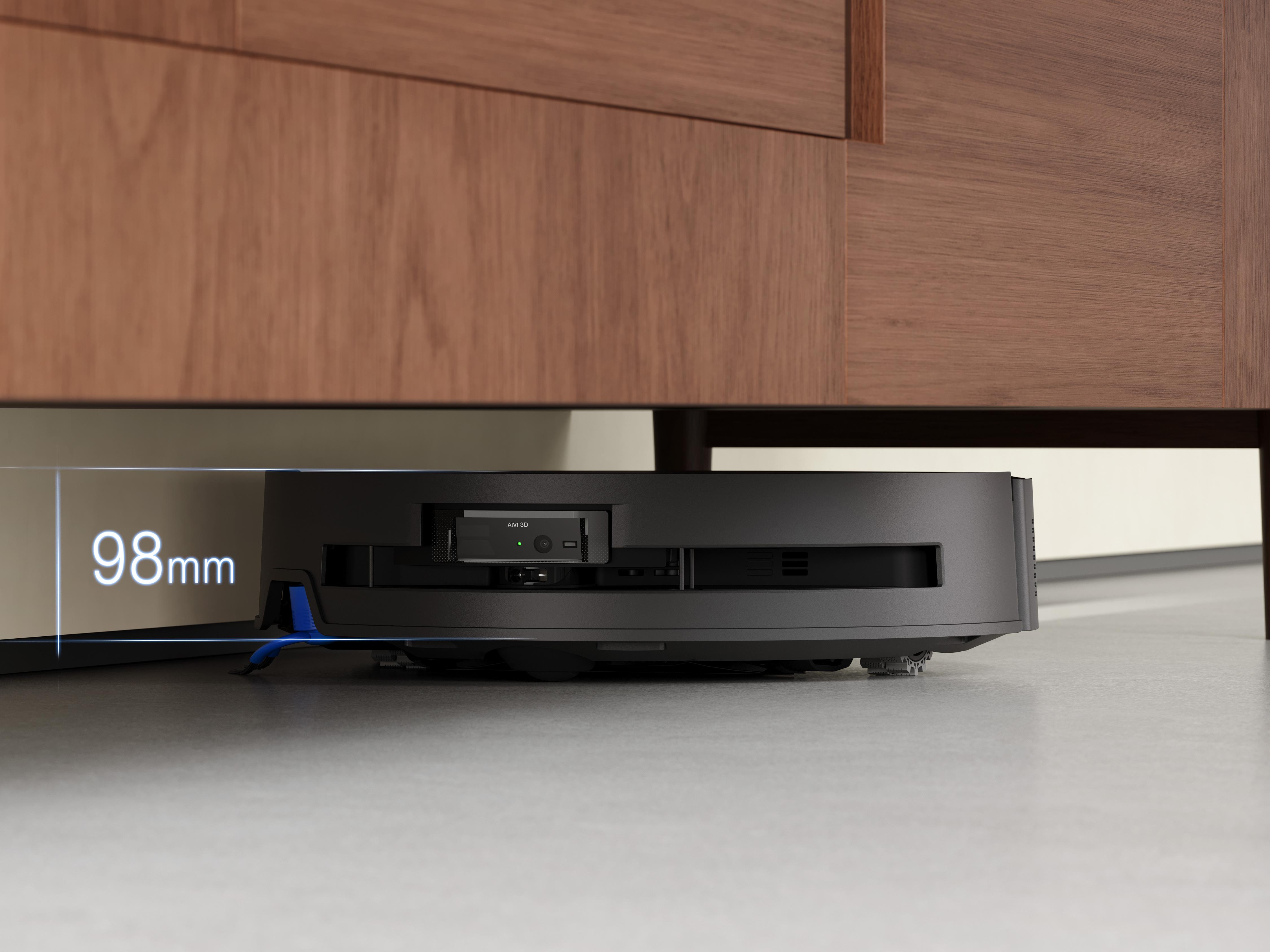 Czarny robot odkurzający pod drewnianą szafką. Wymiar to 98 mm. Widoczna niebieska szczotka.