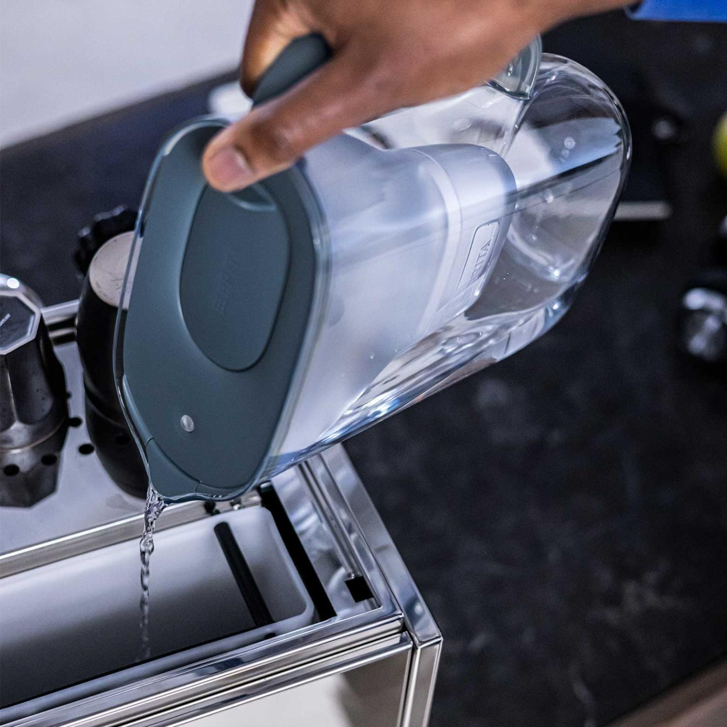 Osoba wlewa wodę z przezroczystego dzbanka z szarym uchwytem do maszyny ze stali nierdzewnej. Woda płynie.
