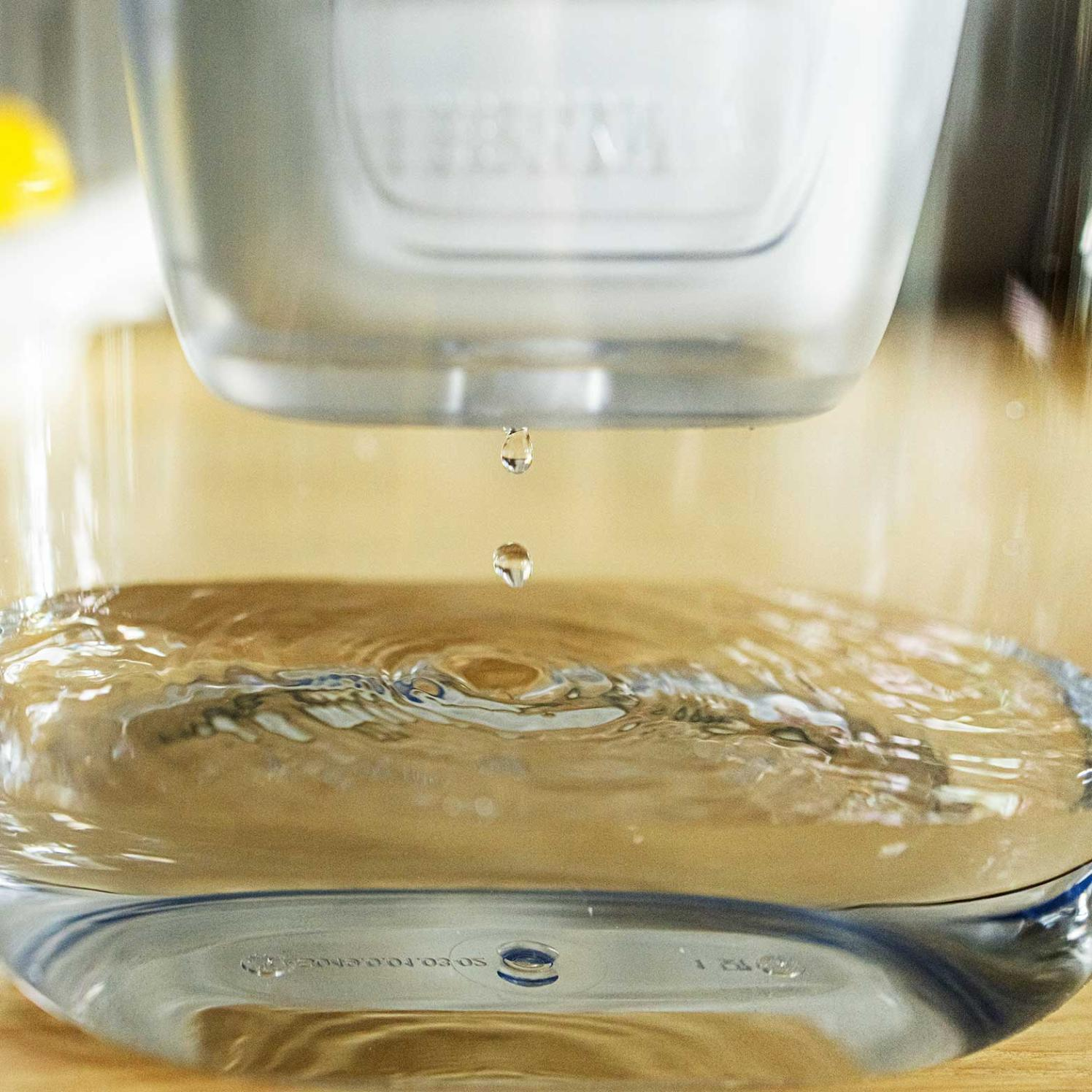 Krople wody spadają z filtra do szklanego pojemnika wypełnionego wodą. Tło to drewniana powierzchnia.