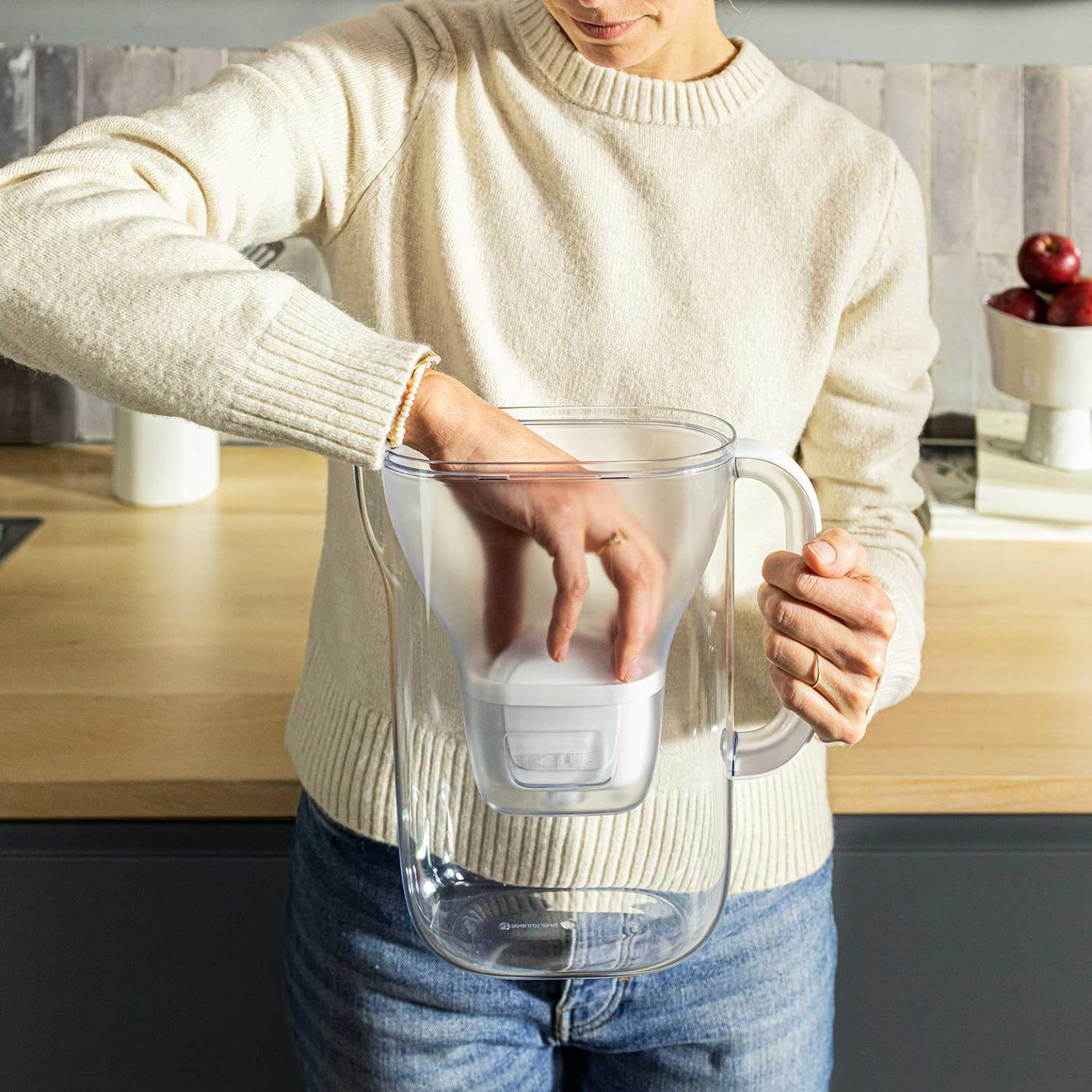 Kobieta w kremowym swetrze i dżinsach wkłada rękę do dzbanka z filtrem do wody. Jabłka w misce w tle.