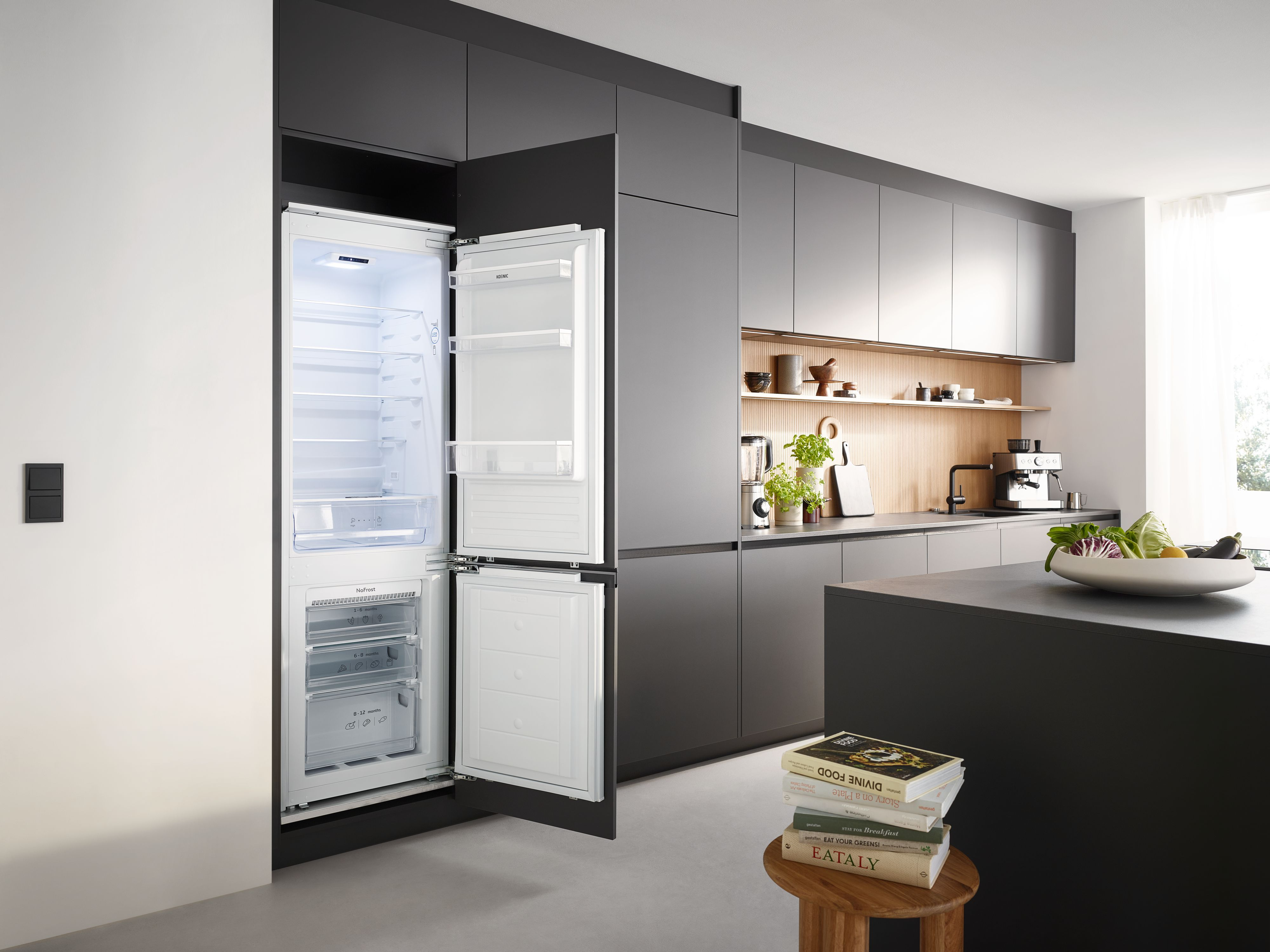 Moderne Küche mit offenem Kühlschrank. Bücher auf einem Hocker, Gemüse auf der Theke. Graue Schränke und weiße Wände.