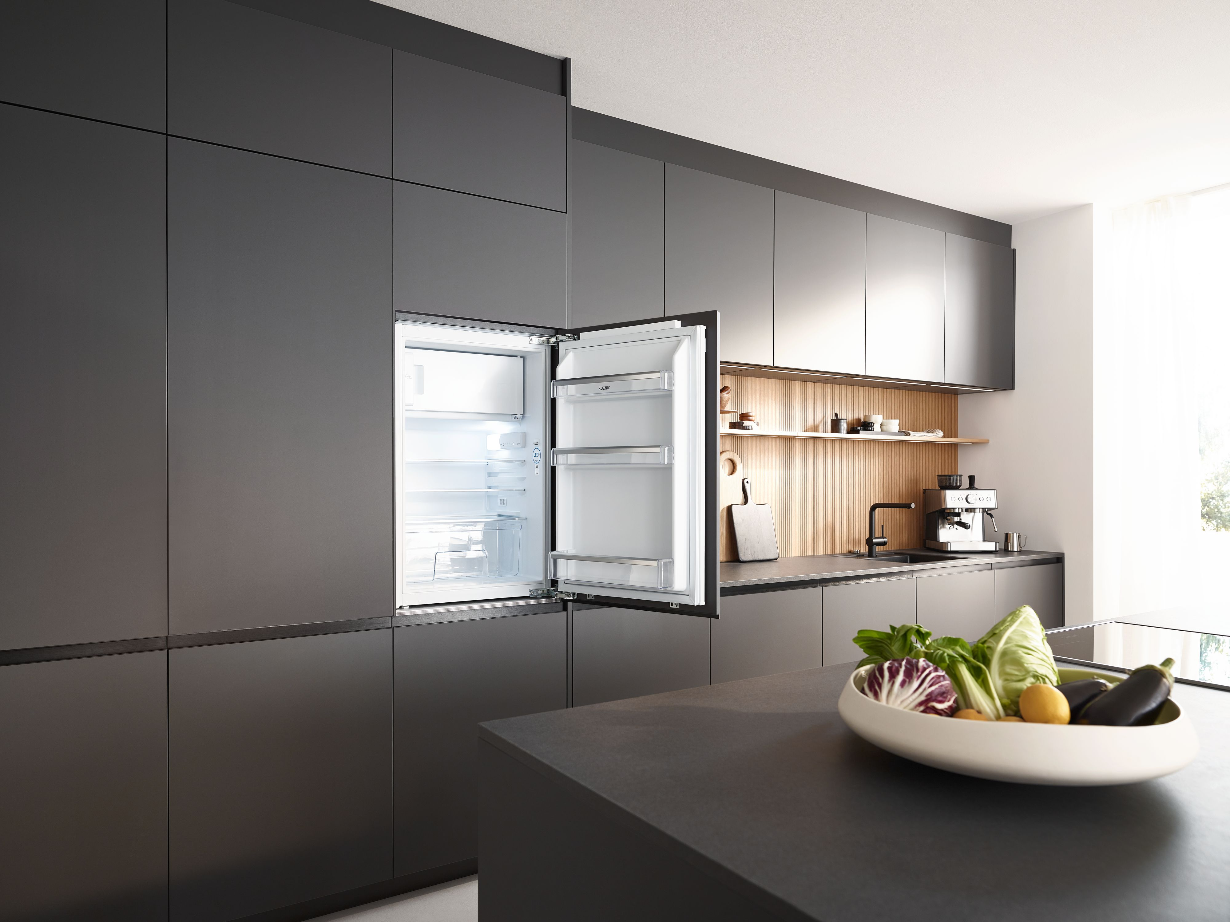 Eine moderne Küche mit geöffnetem Kühlschrank, Obstschale und dunklen Schränken. Eine Kaffeemaschine steht auf der Theke.