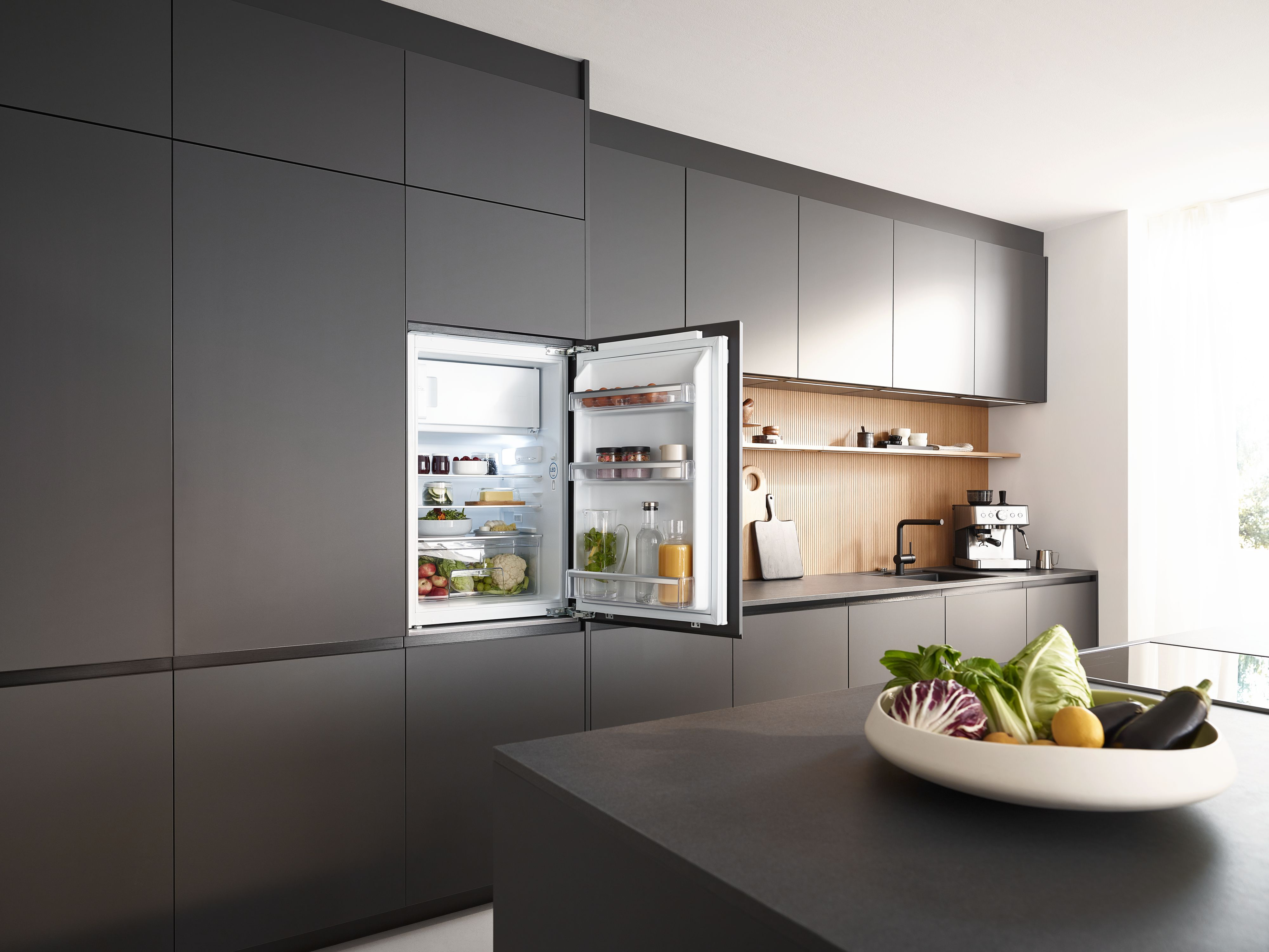 Moderne Küche mit offenem Kühlschrank. Gemüse in einer Schüssel. Schwarze Schränke, Espressomaschine.