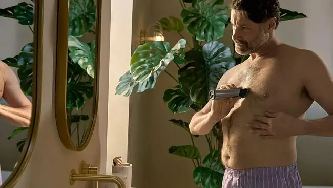 Mężczyzna bez koszuli goli włosy na klatce piersiowej trymerem w łazience. Lustro i zielone rośliny w tle.