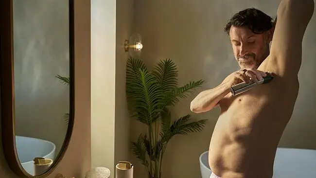 Mężczyzna goli pachy golarką elektryczną w łazience. W tle widoczny jest lustro i roślina.