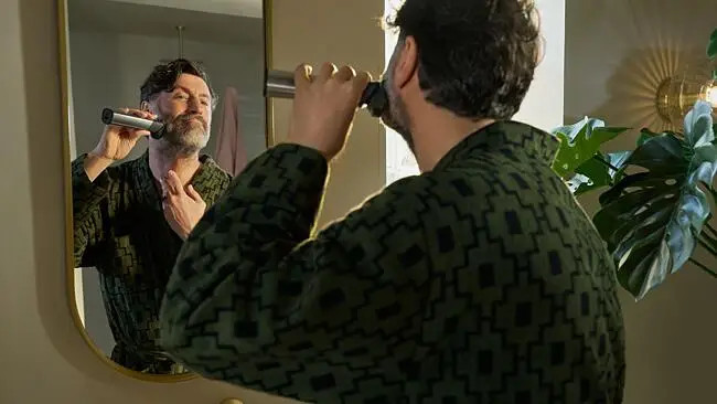 Mężczyzna goli brodę przed lustrem. Ma na sobie zielony szlafrok. Lustro odbija jego twarz i łazienkę.