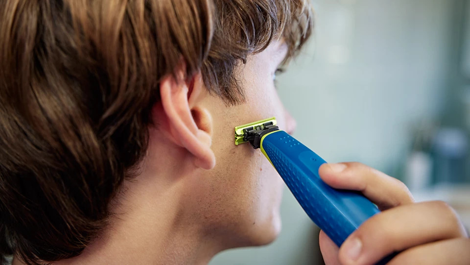 Mężczyzna goli policzek niebiesko-żółtą golarką elektryczną. Jego włosy są brązowe, a skóra jasna. Tło jest rozmyte.