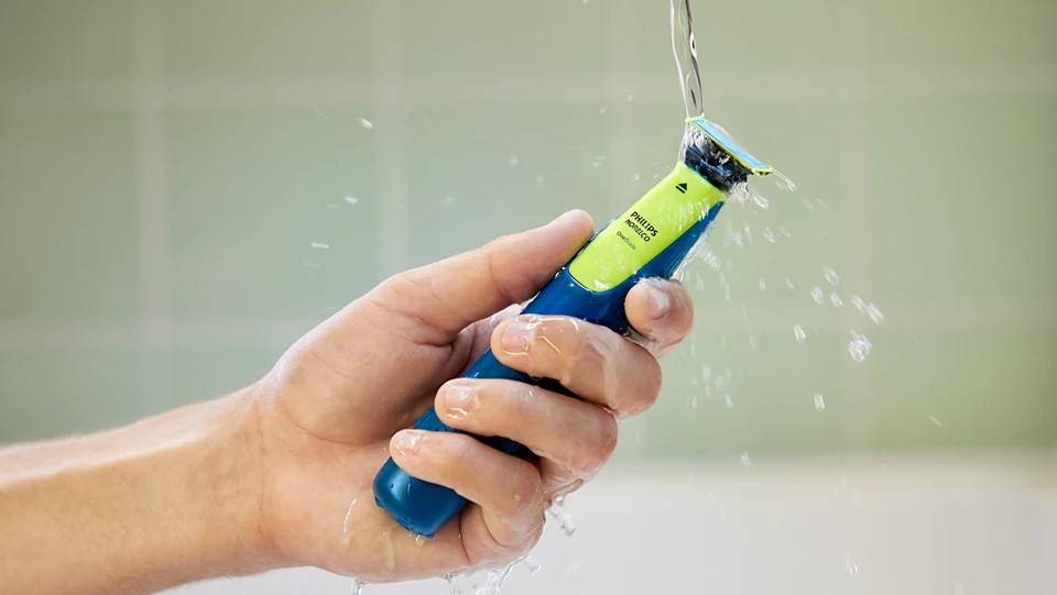 Dłoń trzyma niebiesko-zieloną golarkę Philips Norelco OneBlade pod bieżącą wodą na tle jasnozielonych płytek.