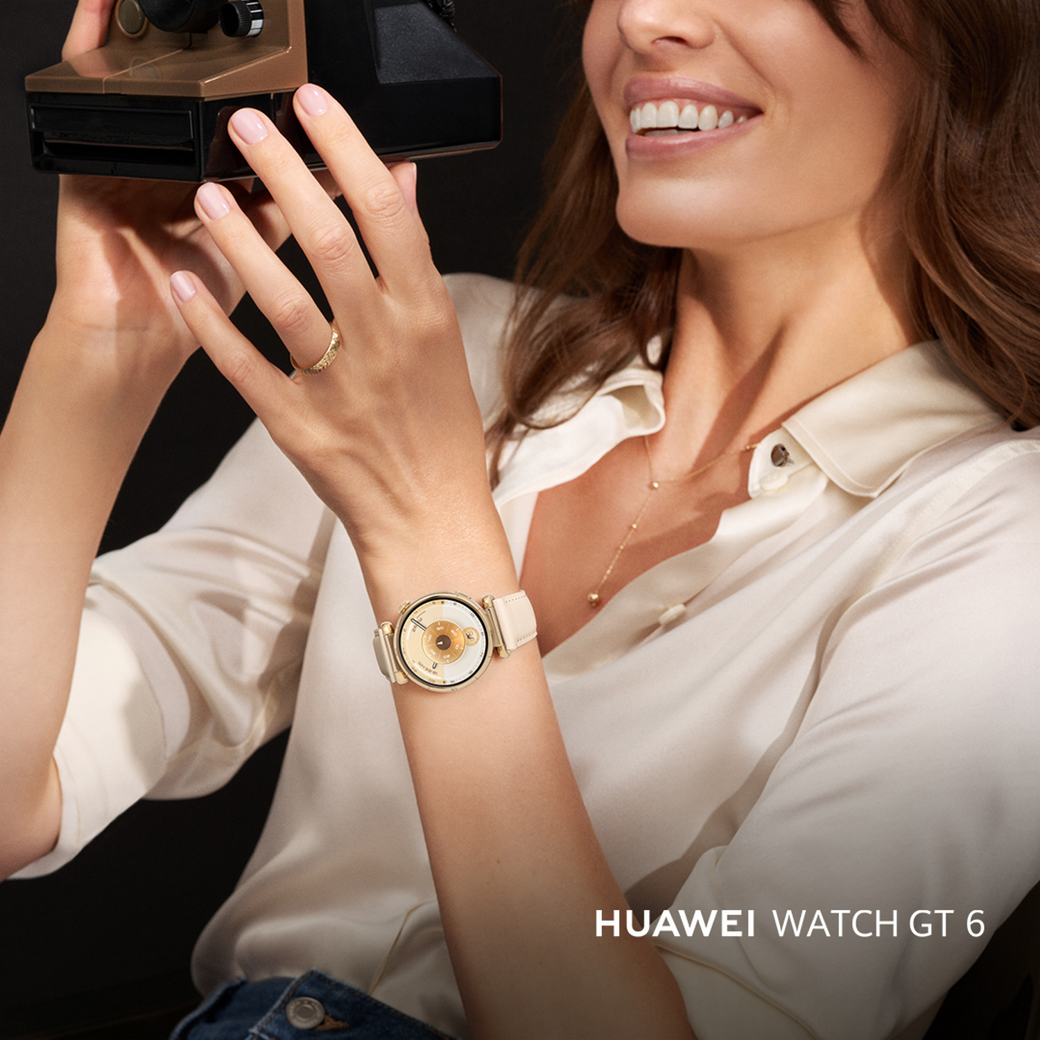 Mujer sonriendo, con reloj, sosteniendo cámara. El reloj tiene una correa crema y esfera dorada. El fondo está borroso.
