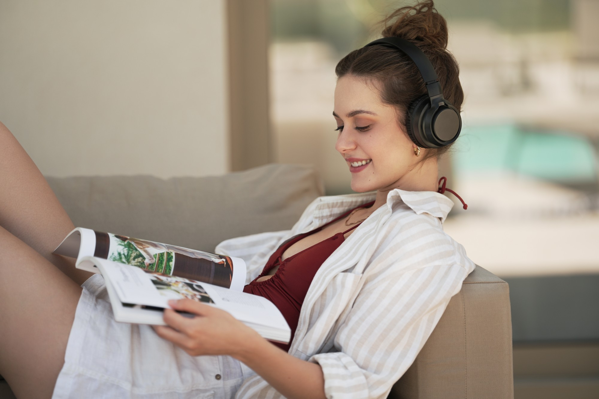 Kobieta w słuchawkach czyta magazyn na kanapie. Uśmiecha się. Ma na sobie czerwony kostium kąpielowy i koszulę w paski.