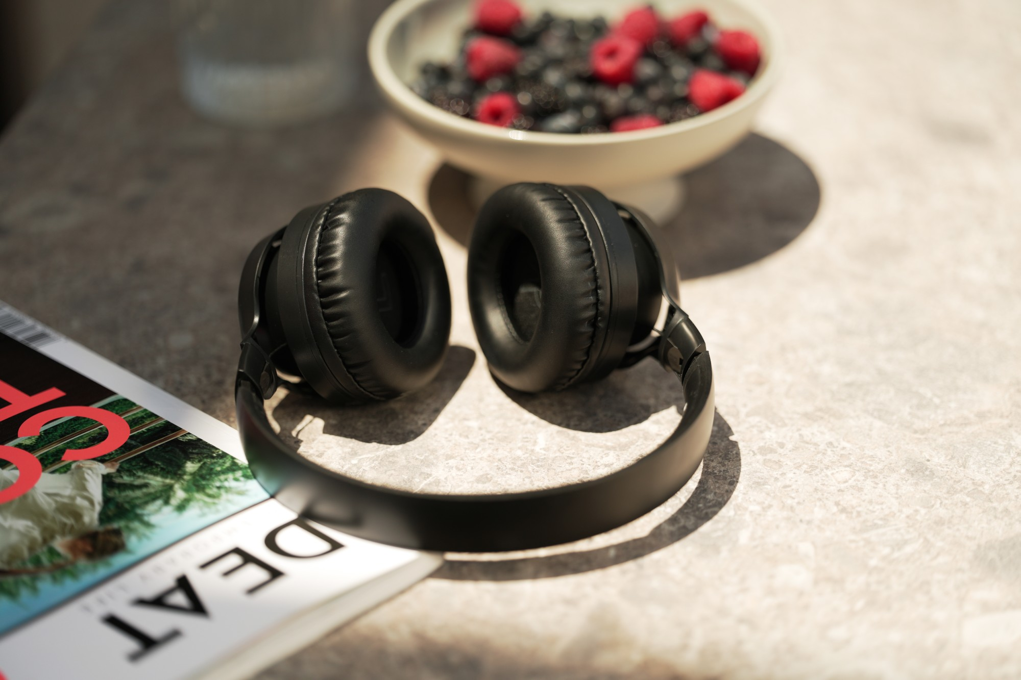 Czarne słuchawki leżą na magazynie. Miska mieszanych jagód z tyłu. Scena oświetlona miękkim światłem słonecznym.