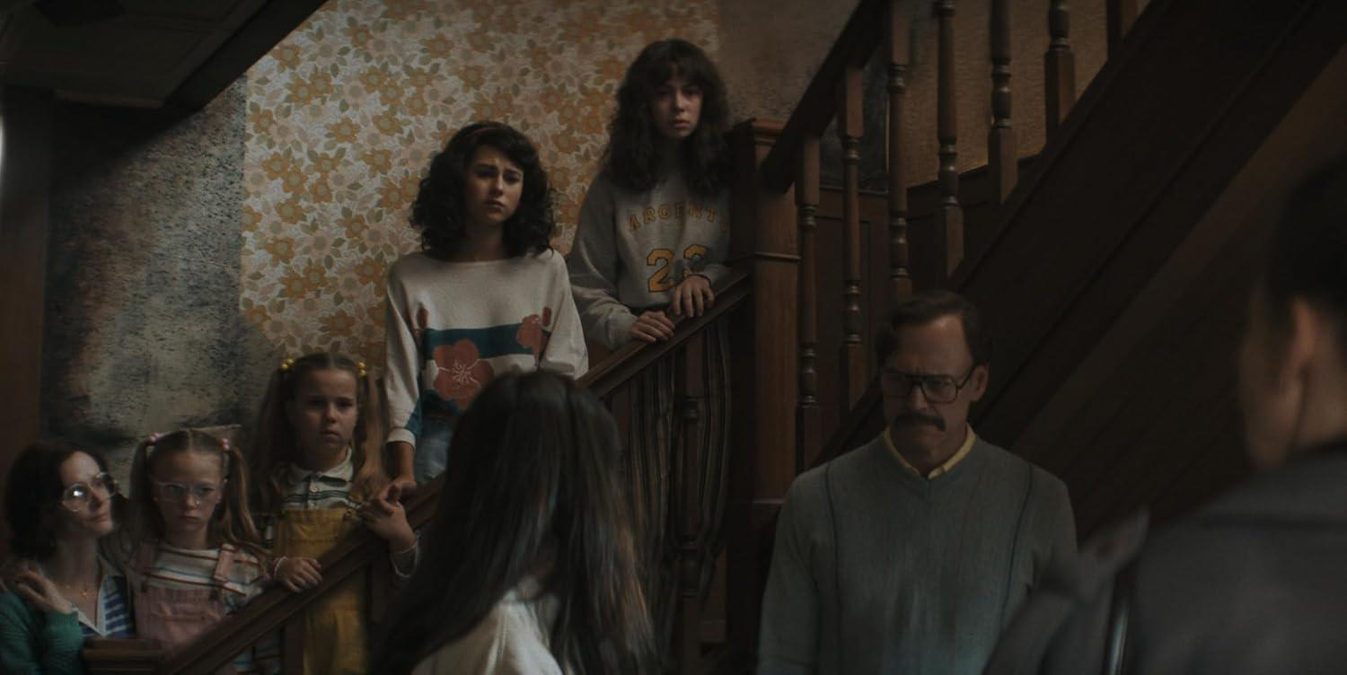 Eine Familie steht auf einer Treppe. Frauen und Kinder stehen im Vordergrund. Ein Mann mit Brille steht unten. Blumige Tapete ist sichtbar.
