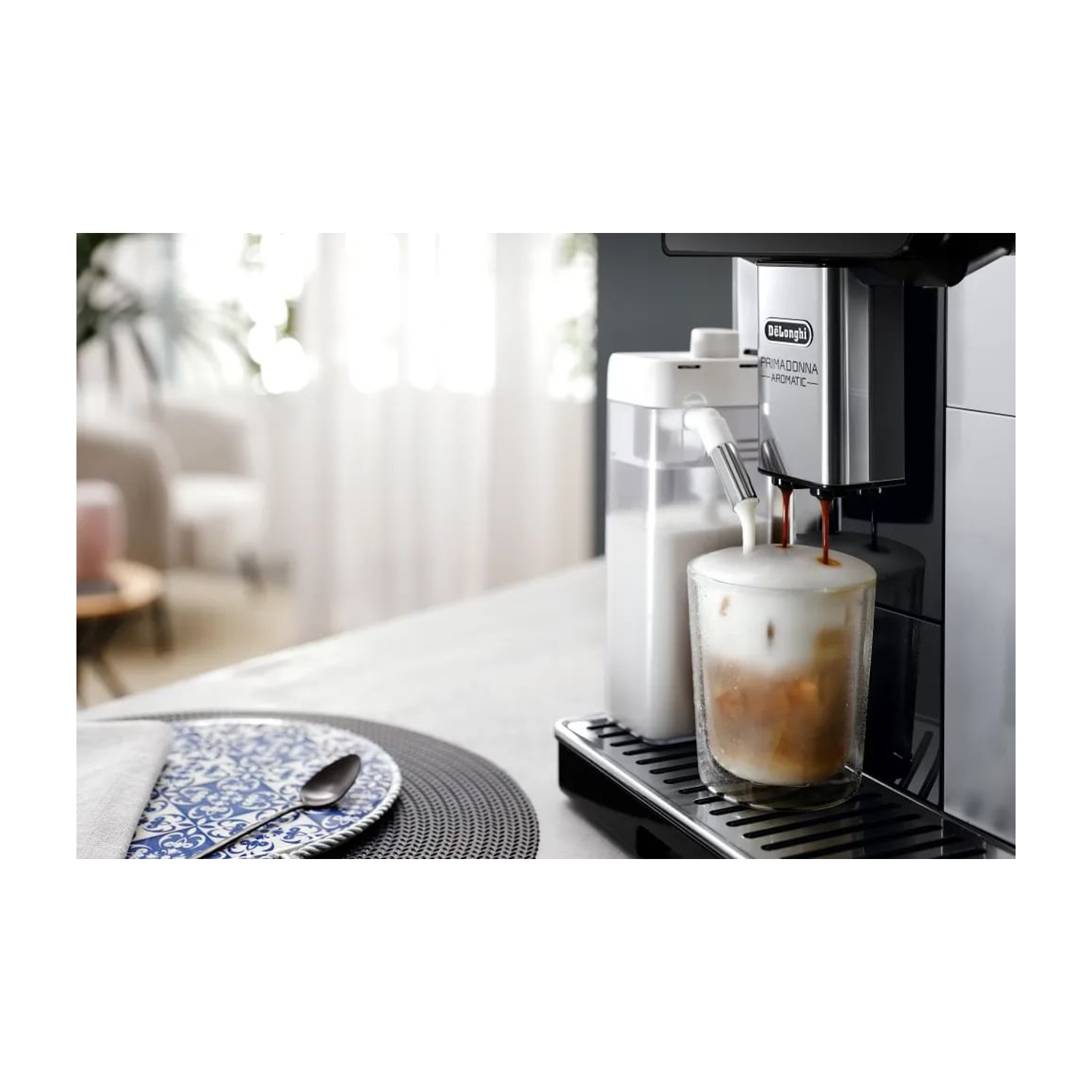 Macchina da caffè che eroga caffè in un bicchiere. Contenitore del latte a sinistra. Piatto e cucchiaio sul tavolo.