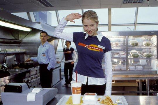 Eine Frau in einer Fast-Food-Uniform salutiert. Zwei weitere Mitarbeiter sind im Hintergrund. Essen und eine Kasse sind vorne.