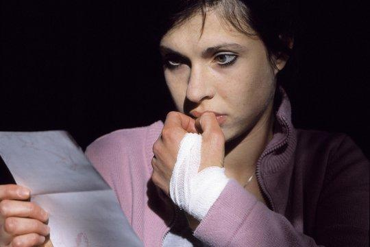 Eine Frau in einer rosa Jacke hält ein Papier und beißt sich die Nägel. Ihre bandagierte Hand ruht an ihrem Mund. Dunkler Hintergrund.