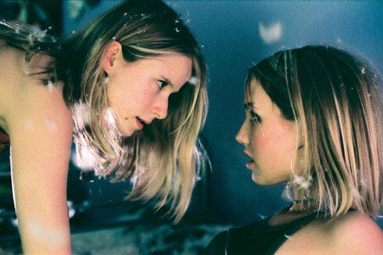 Zwei Frauen stehen sich gegenüber, Federn im Haar. Eine blickt nach unten, die andere schaut nach vorn. Dunkle Kleidung und blauer Hintergrund.