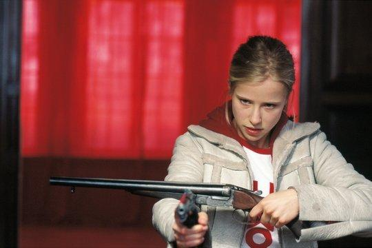Eine Frau hält eine Schrotflinte und zielt auf den Betrachter. Sie trägt eine weiße Jacke, ein rotes Oberteil und hat blondes Haar. Rote Vorhänge sind im Hintergrund.