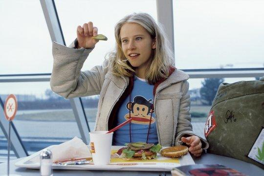 Blondes Mädchen hält eine Gurke über einem Burger. Sie trägt ein Affen-Shirt und eine Jacke. Burger, Getränk und Tasche auf dem Tisch.
