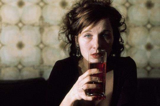 Eine Frau hält ein Glas rote Flüssigkeit und schaut den Betrachter an. Sie hat dunkle Haare und trägt ein schwarzes Oberteil. Der Hintergrund ist gemustert.