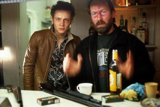 Zwei Männer stehen hinter einer Theke mit einer Schrotflinte, Tassen und Flaschen. Einer trägt eine Lederjacke, der andere eine schwarze Jacke.