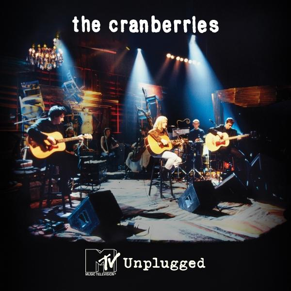 The Cranberries unplugged. Bandmitglieder mit Gitarren auf der Bühne. Bühne hat Stühle und Lichter. MTV Unplugged Logo ist sichtbar.