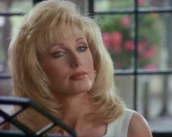 Eine Frau mit blondem Haar und blauen Augen blickt zur Seite. Sie trägt ein weißes Oberteil. Im Hintergrund befindet sich ein Fenster mit Blumen.