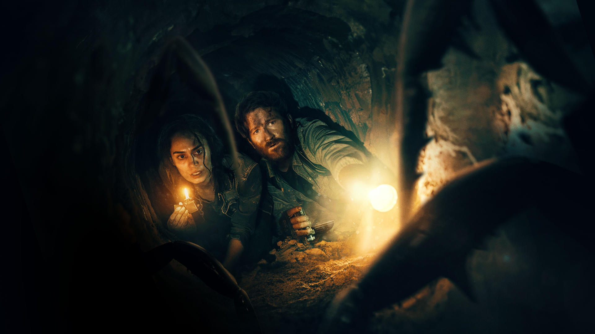 Zwei Menschen kriechen durch einen dunklen Tunnel, eine mit einer Kerze, der andere mit einer Taschenlampe.