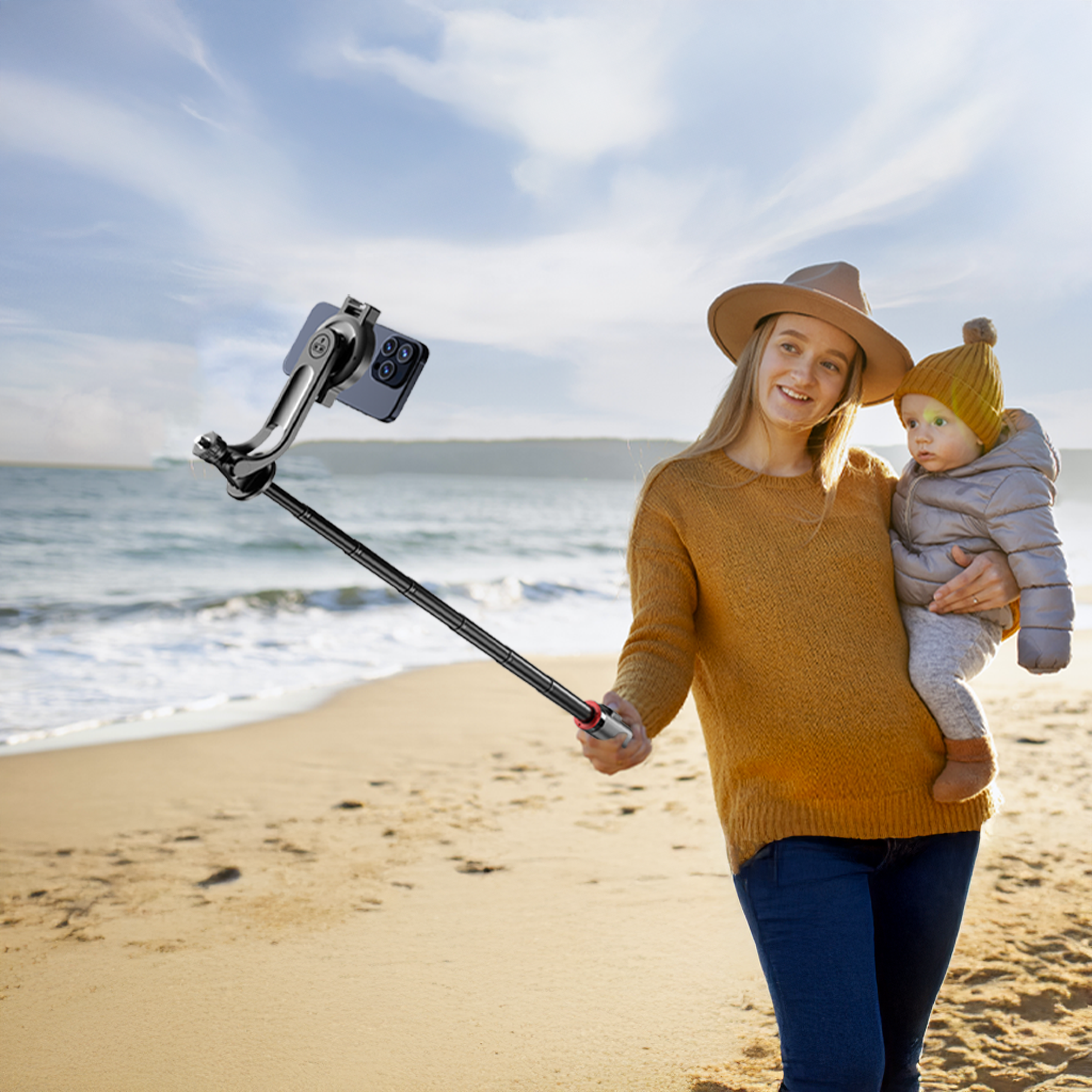 Eine Frau hält ein Baby am Strand und macht ein Selfie mit einem Telefon, das an einem Selfie-Stick befestigt ist. Meer und Himmel im Hintergrund.