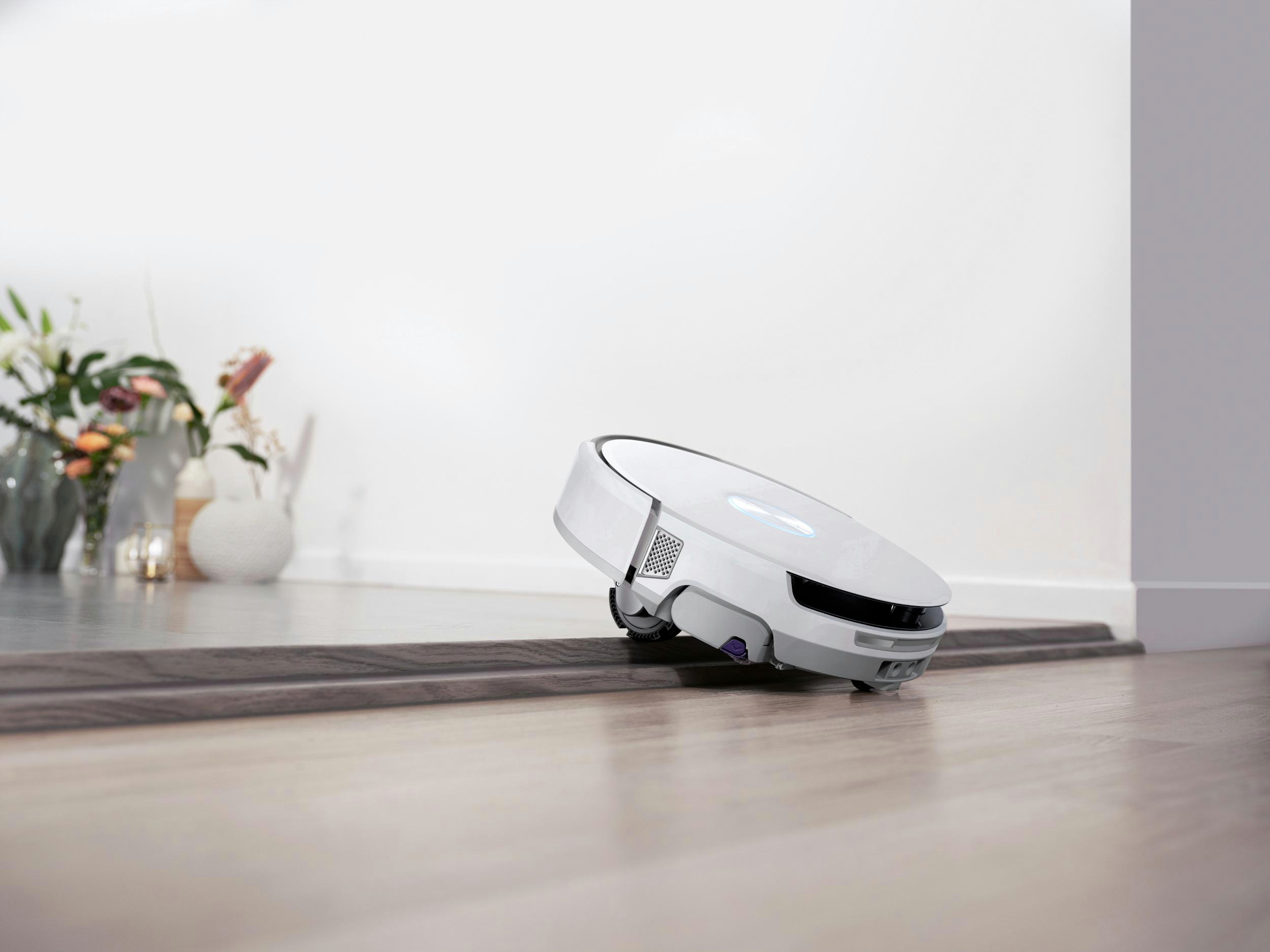 Ein weißer Roboter-Staubsauger auf einem Holzboden. Im Hintergrund eine Vase mit Blumen. Die Wand ist weiß.