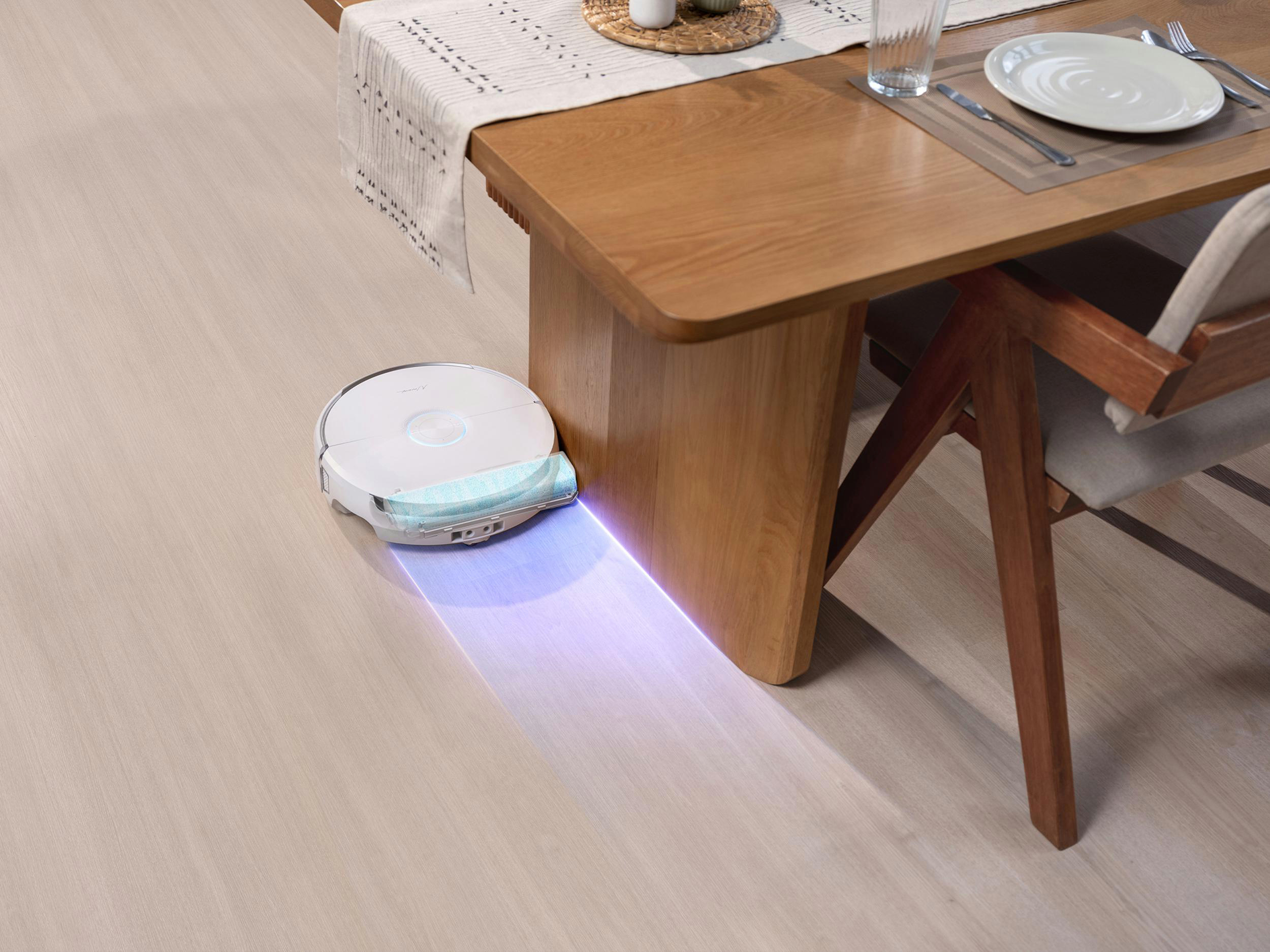 Ein Roboter-Staubsauger reinigt unter einem Holztisch, der für eine Mahlzeit gedeckt ist. Der Boden ist helles Holz.