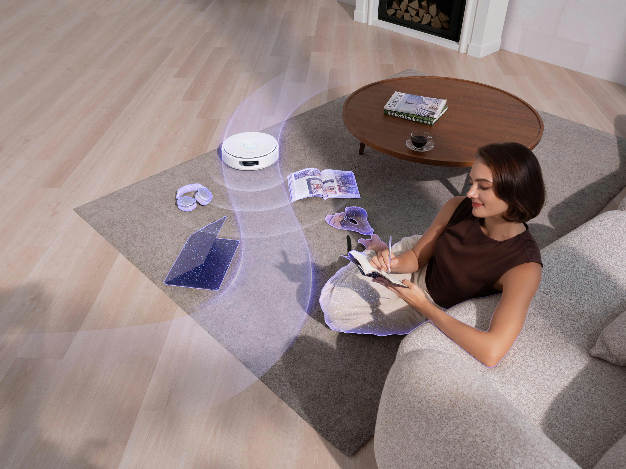 Eine Frau sitzt auf einem Sofa und schreibt in ein Notizbuch. Ein Roboterstaubsauger reinigt den Boden. Ein Couchtisch ist neben ihr.