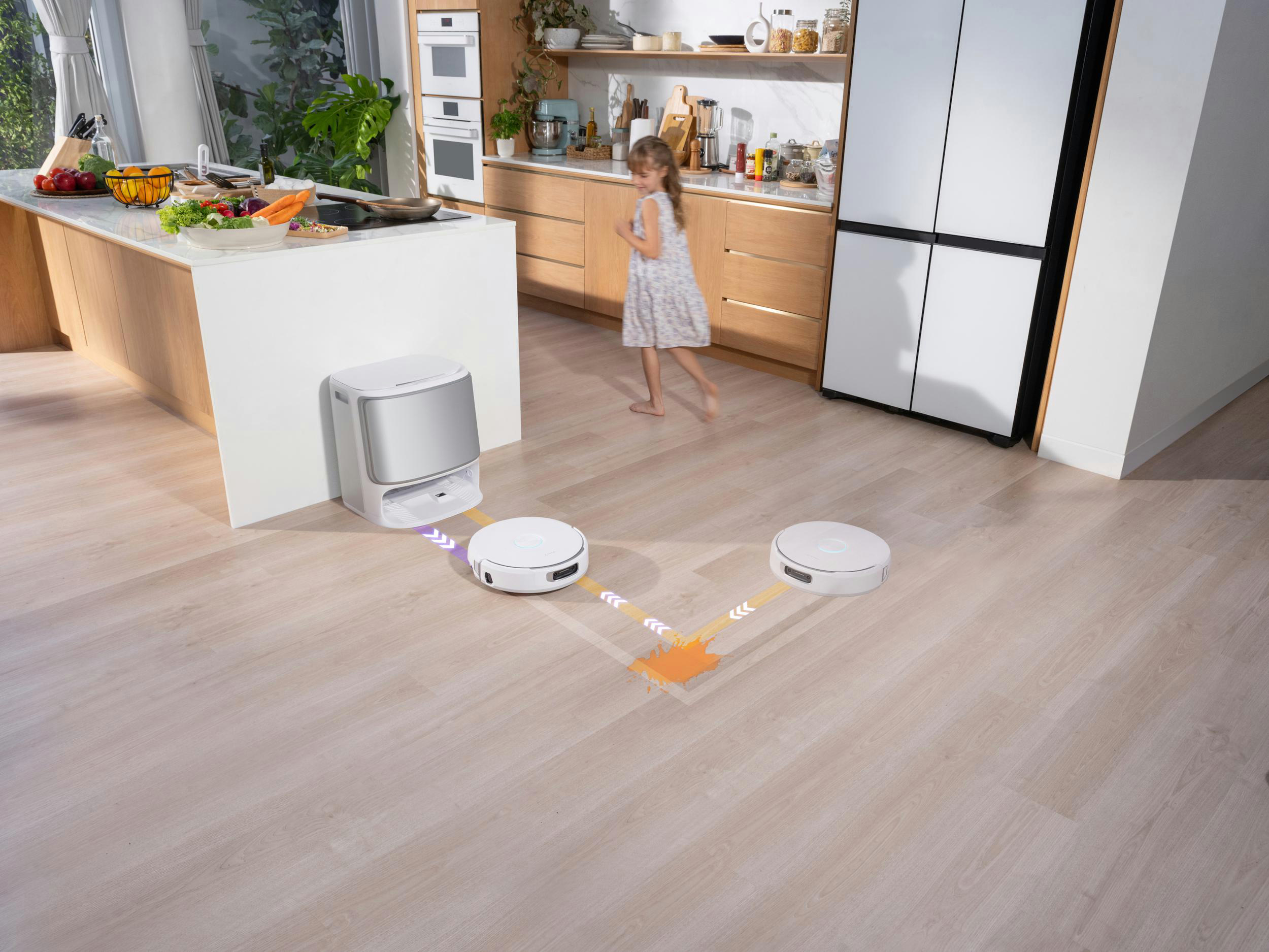 Eine Küchenszene mit einem rennenden Mädchen. Zwei Roboterstaubsauger reinigen den Boden. Eine weiße Kücheninsel enthält Lebensmittel.