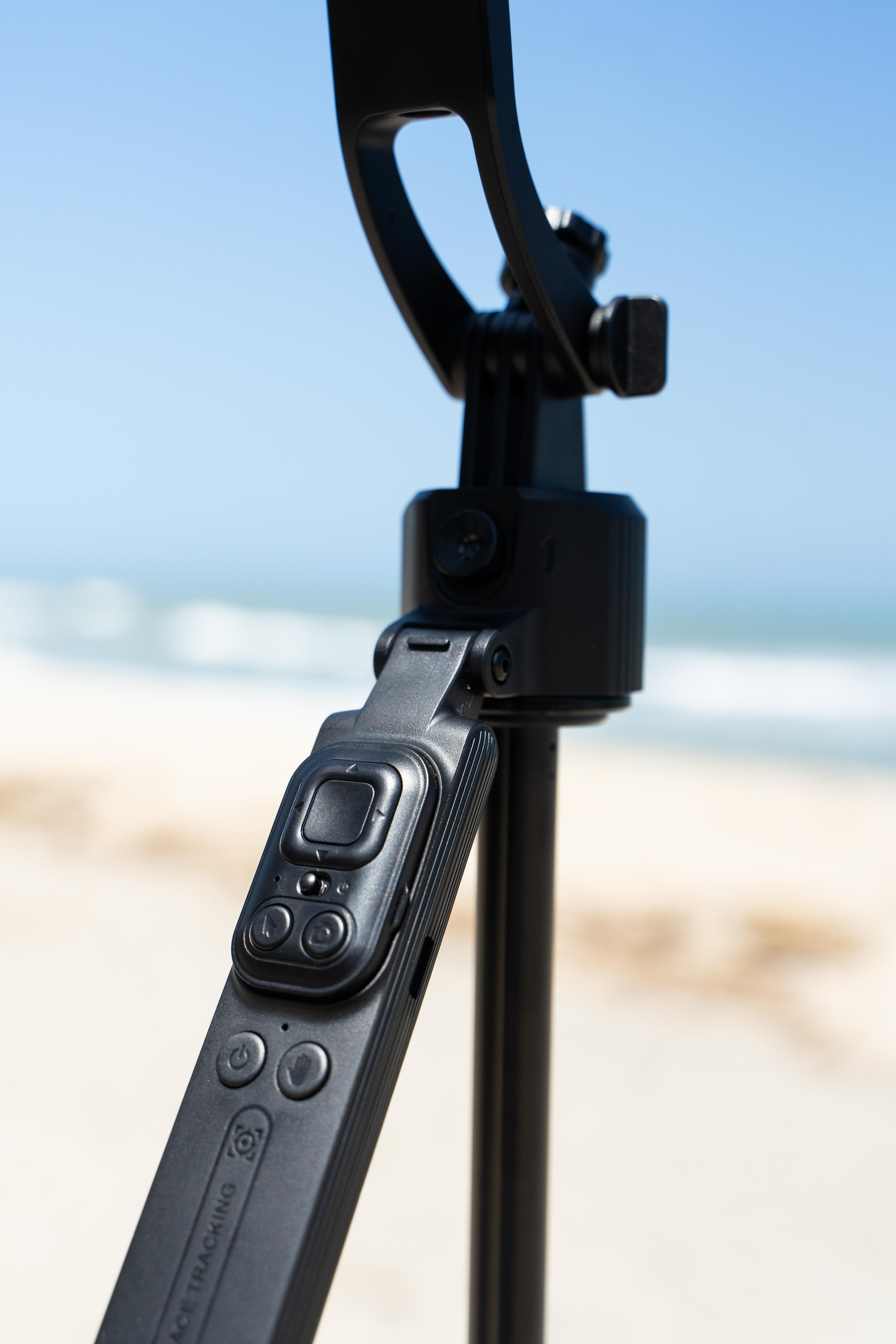 Schwarzer Kamerastabilisator am Strand mit blauem Himmel und Ozean im Hintergrund.