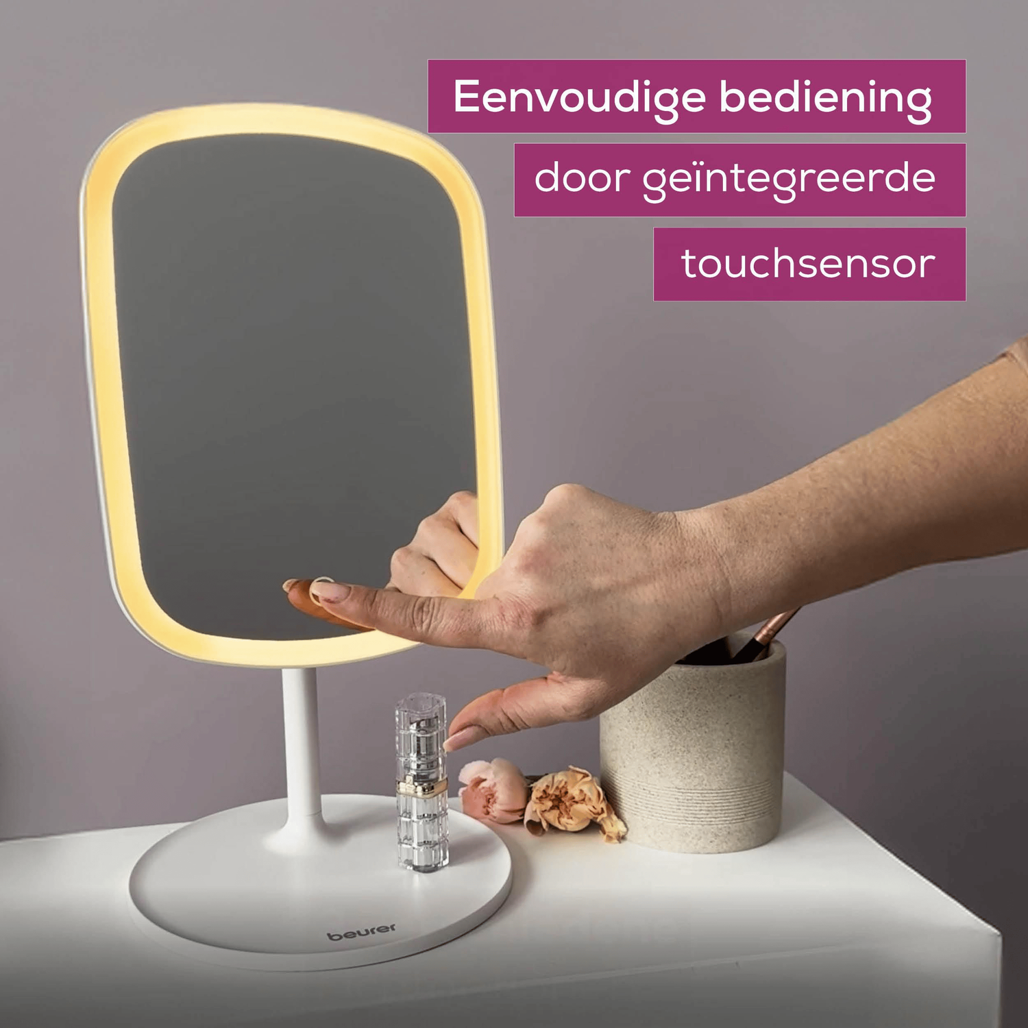 A hand touches a lighted mirror. Other items include a stand, a makeup brush holder, and flowers on a white surface.