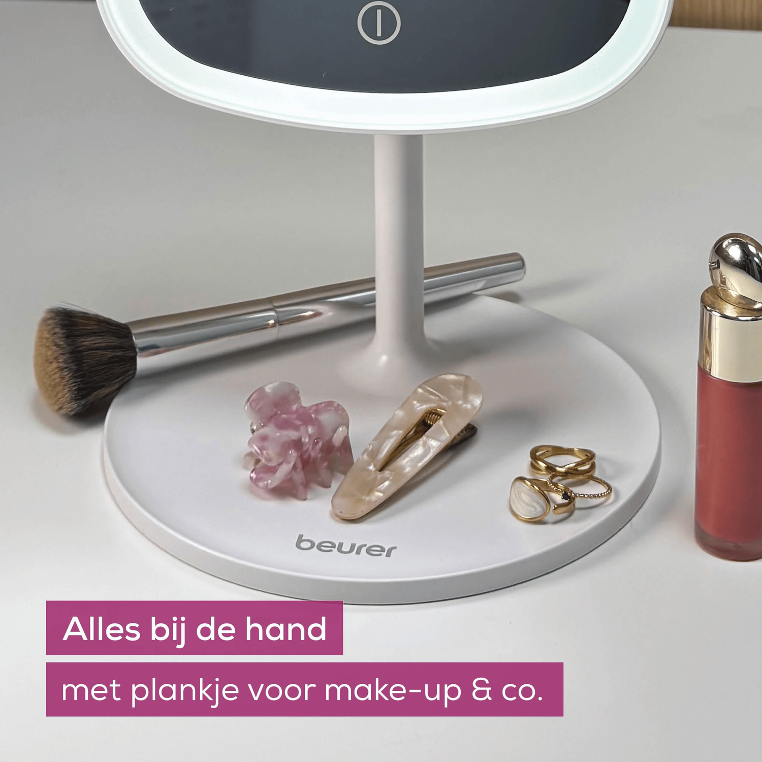 A makeup mirror with accessories: brush, hair clips, rings and lipstick on a white surface.