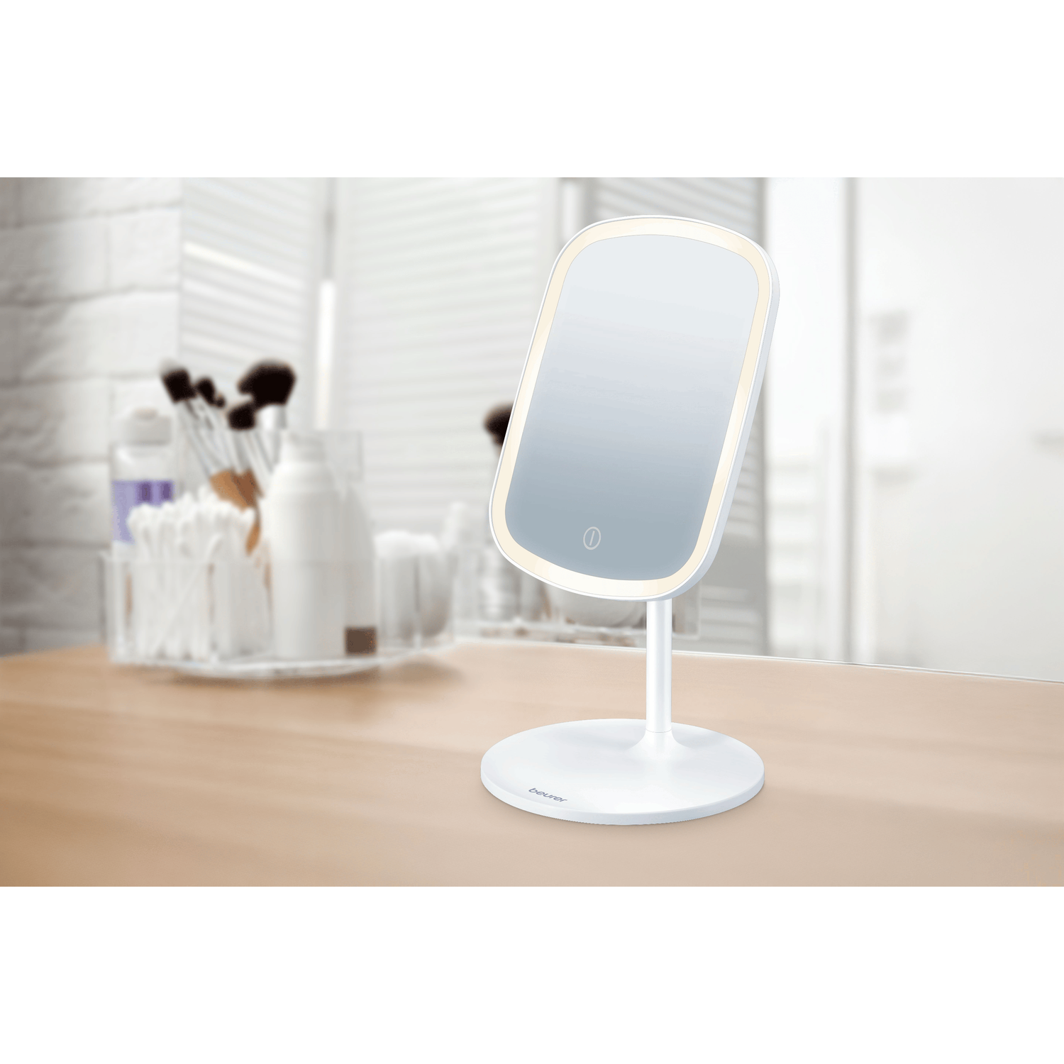A white, illuminated makeup mirror stands on a wooden surface. Background has toiletries.