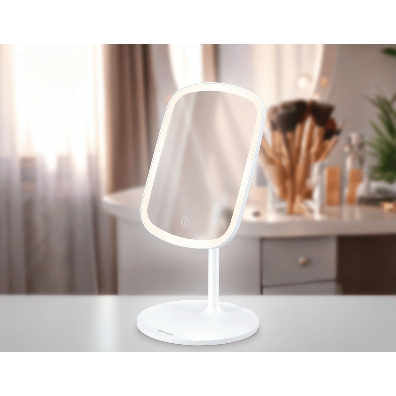 A white illuminated makeup mirror stands on a white table. Soft focus background shows a vanity with brushes and a window.