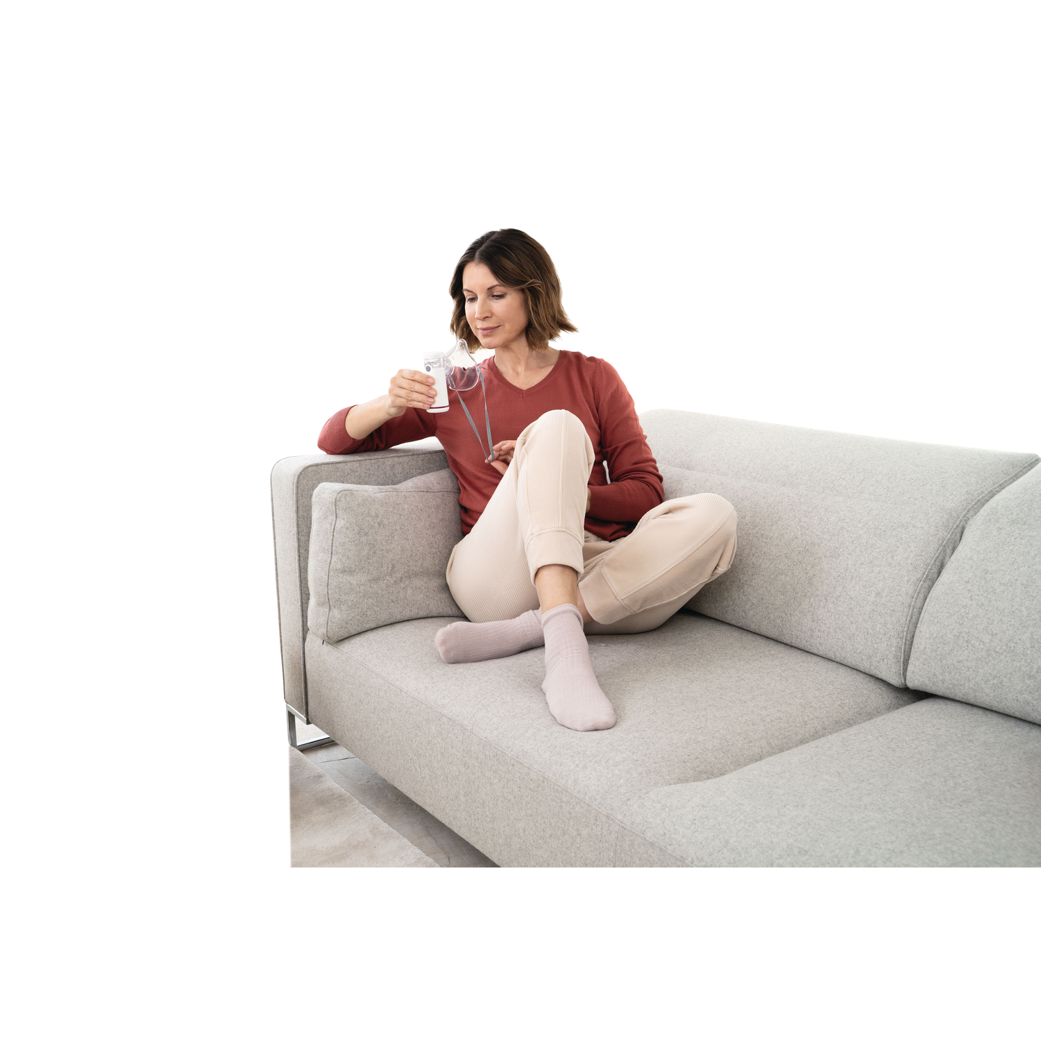 A woman sits on a grey couch, holding a nebulizer. She wears a red shirt, beige pants, and pink socks. The background is white.