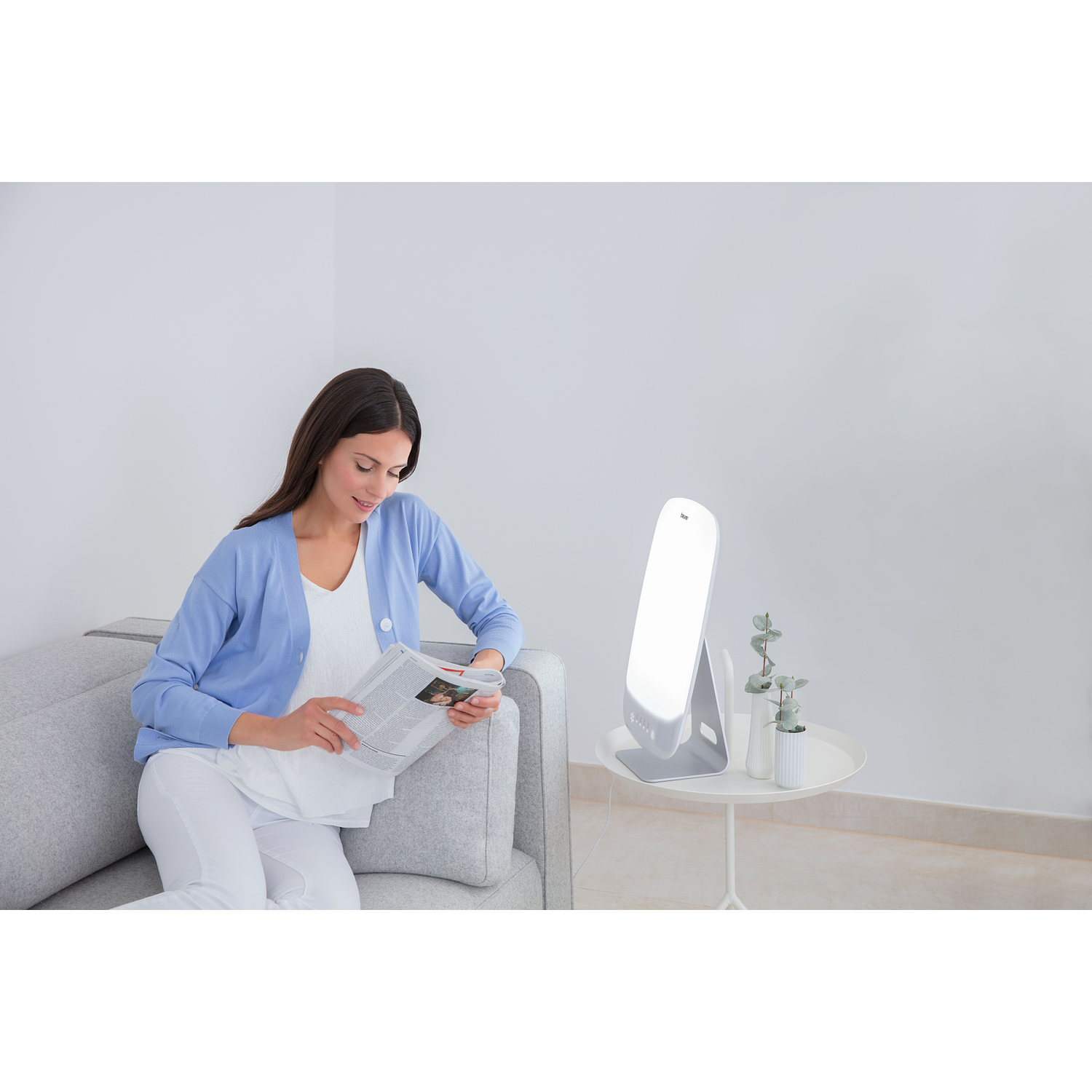 Vrouw op een bank leest een tijdschrift. Een lichttherapielamp en planten op een bijzettafel. De vrouw draagt blauw en wit.