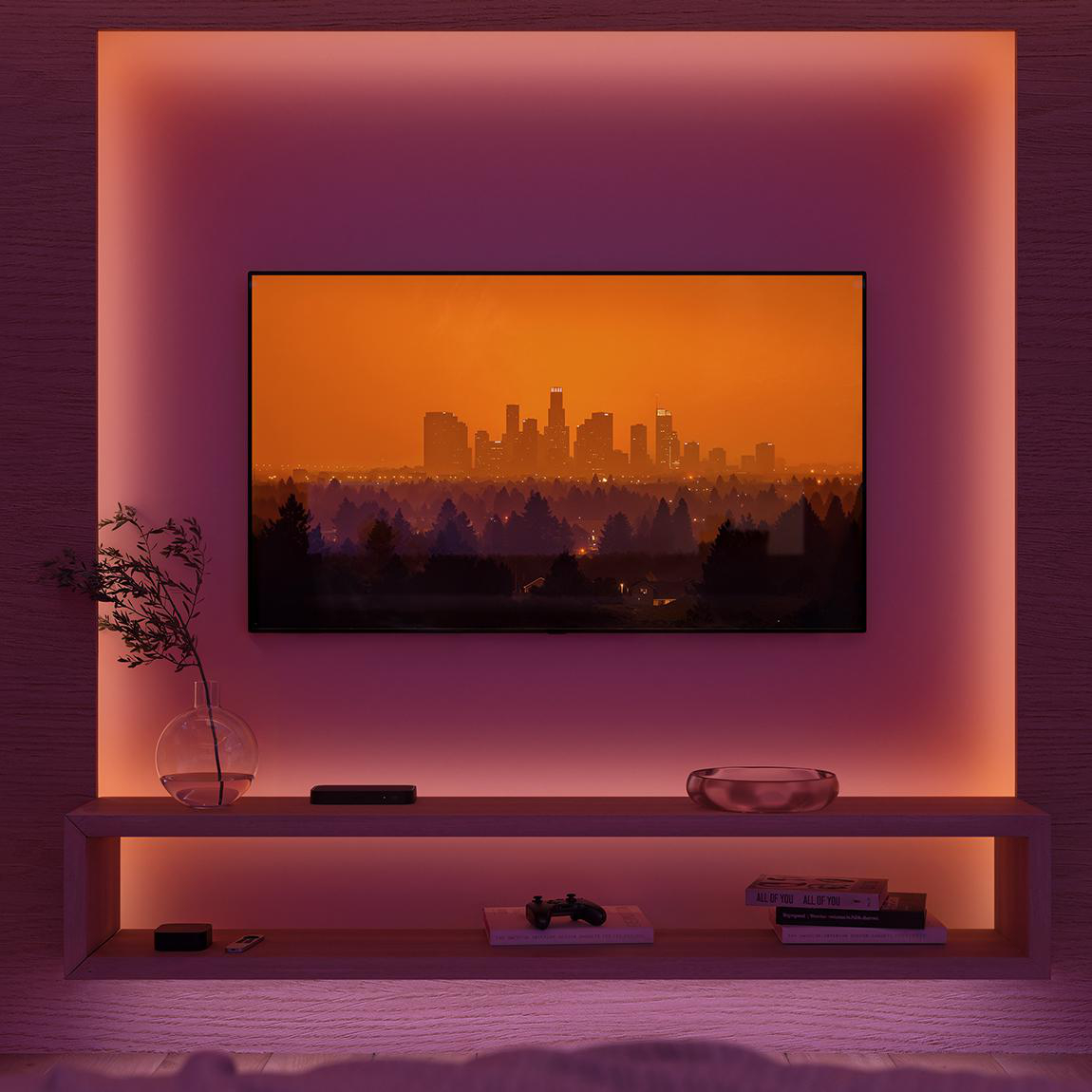 Ein Fernseher zeigt eine Skyline vor einem orangefarbenen Himmel. Ein Regal darunter enthält Dekoration. Rosa Umgebungslicht umrahmt den Fernseher.