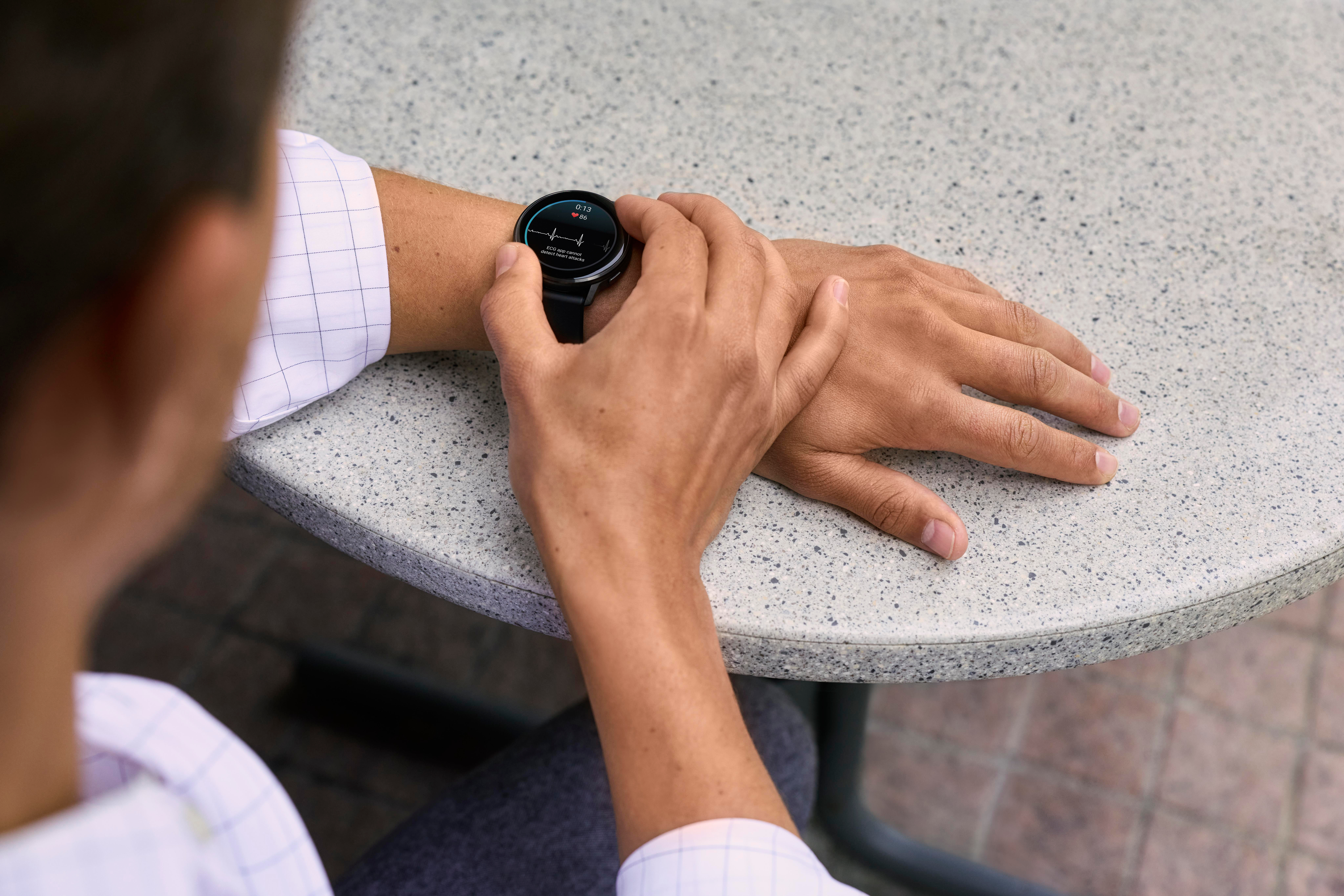Person trägt Smartwatch am Handgelenk und checkt den Bildschirm. Hand liegt auf strukturiertem Tisch. Ziegelmuster im Hintergrund.