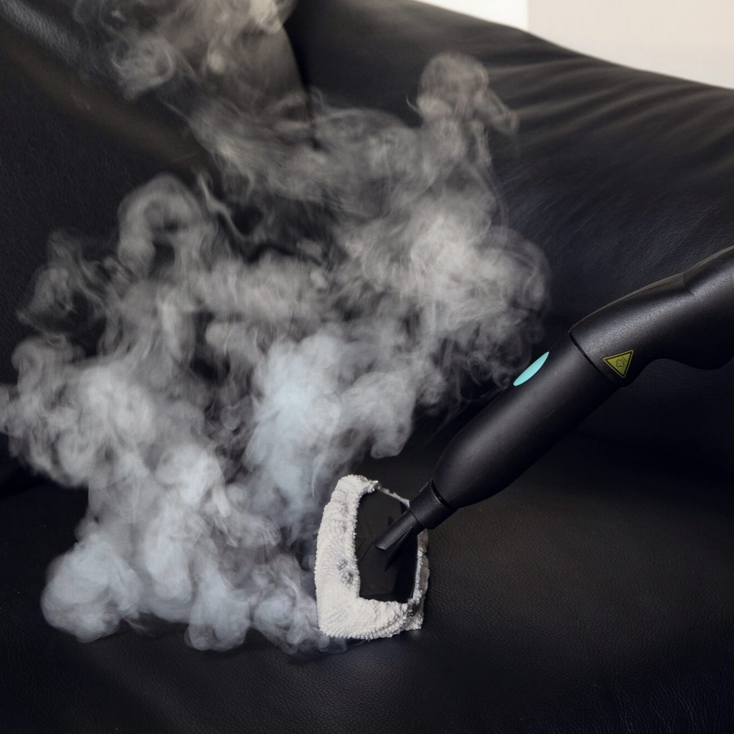Un divano in pelle nera viene pulito con un pulitore a vapore. Il vapore bianco sale dal pulitore, mostrando il processo di pulizia.