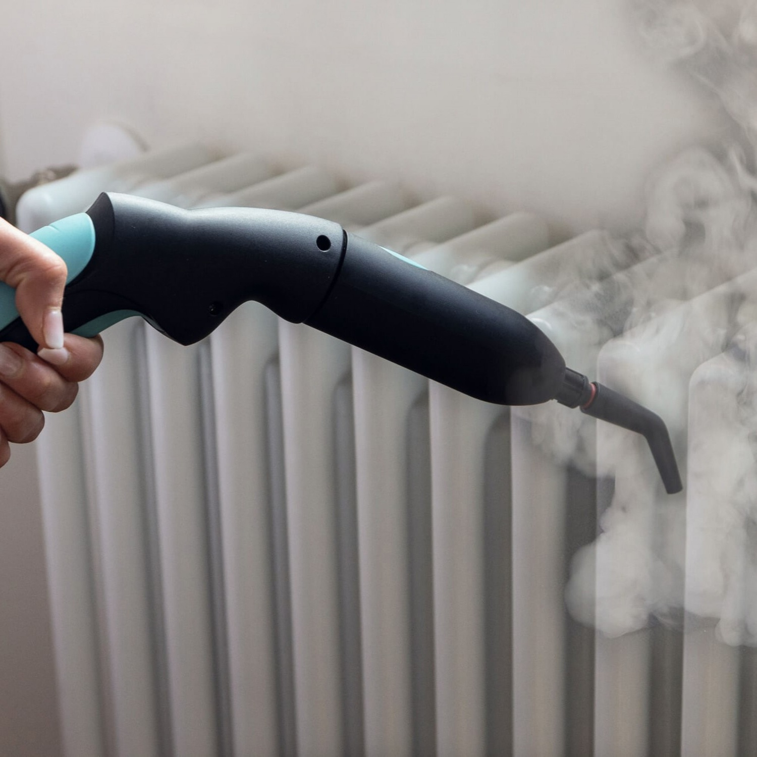 Una mano tiene un pulitore a vapore nero e verde acqua, che emette vapore su un radiatore bianco.