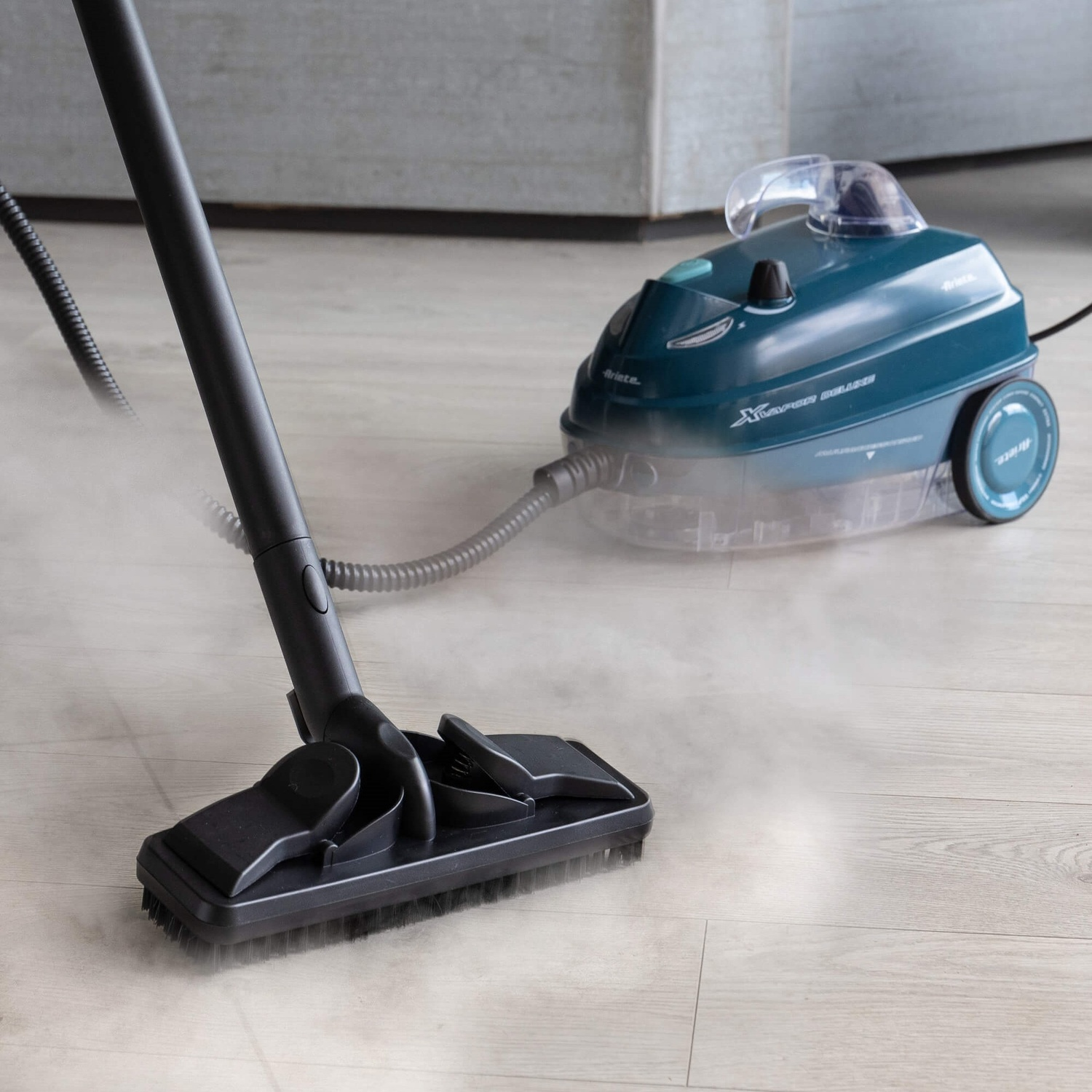 Un pulitore a vapore turchese sta pulendo un pavimento in legno chiaro. Il vapore sale dalla testina del pulitore. Un altro pulitore a vapore è sullo sfondo.