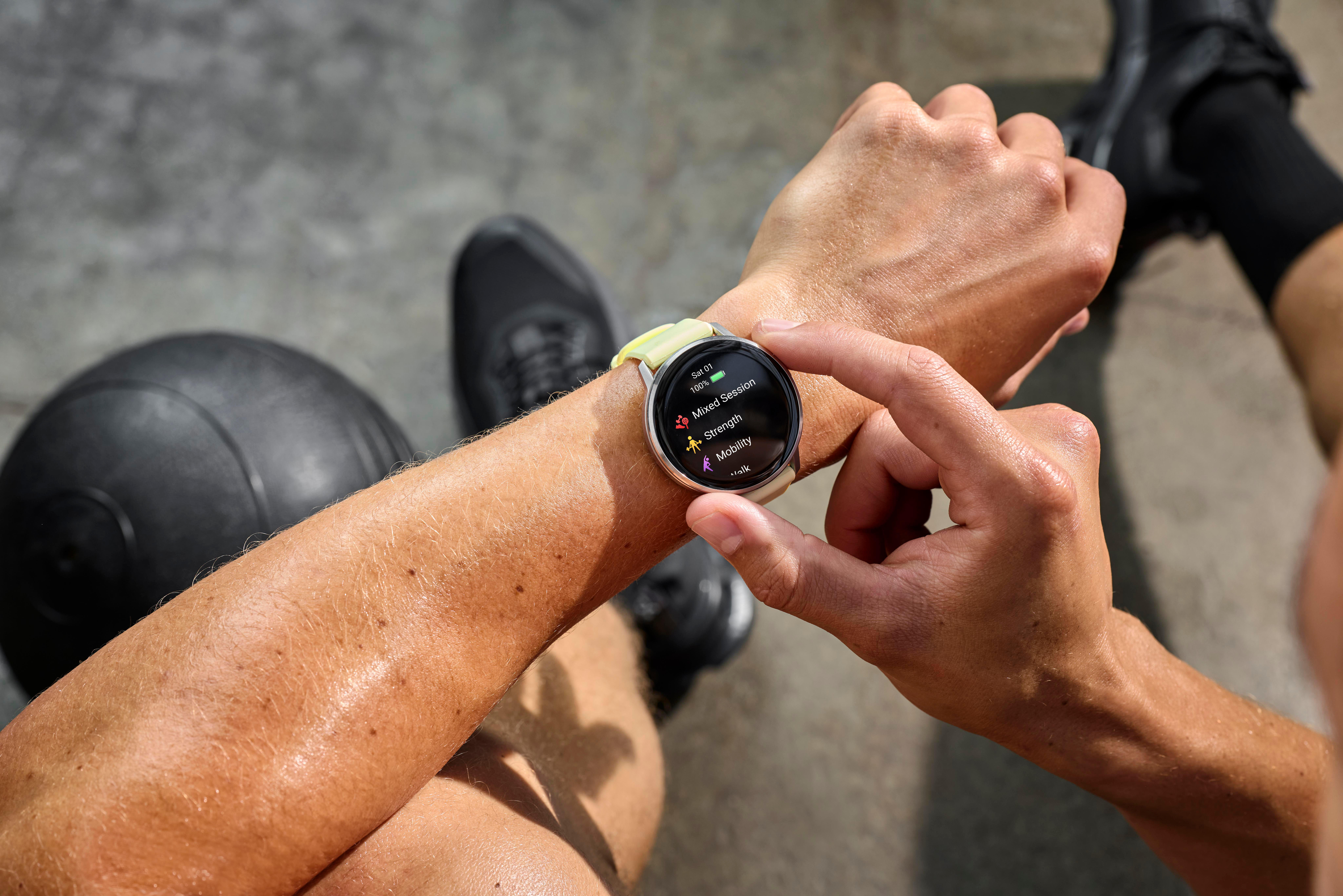 Eine Person, die eine Smartwatch trägt und Workout-Optionen anzeigt. Im Hintergrund befinden sich ein schwarzer Ball und schwarze Schuhe. Die Person berührt die Uhr.