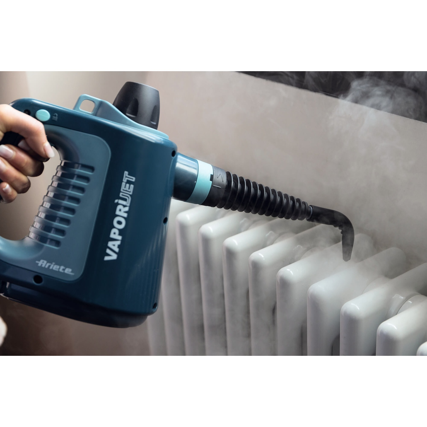 Una mano tiene un vaporizzatore VAPORJET turchese, che pulisce un radiatore bianco. Il vapore viene emesso, pulendo il radiatore.