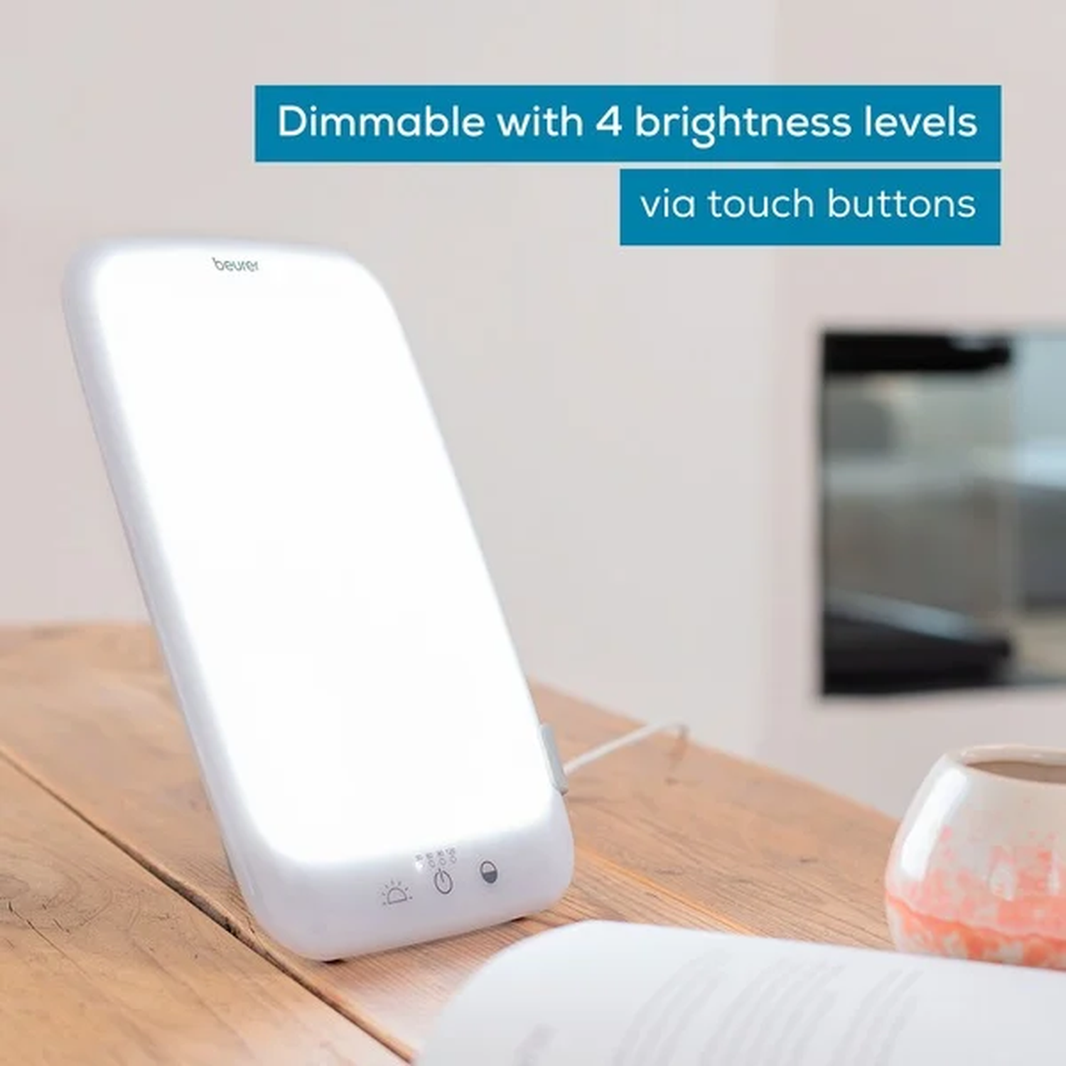 A white therapy lamp on a wooden table. It has touch buttons and four brightness levels. A book and a vase are also on the table.