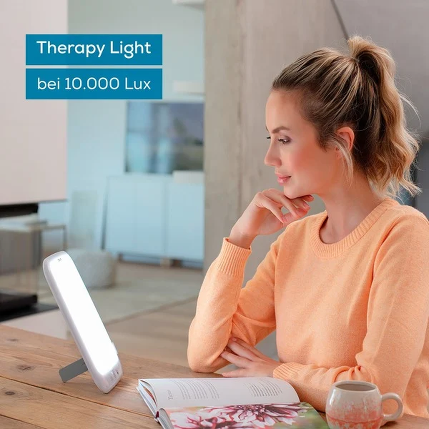 Woman with fair skin looking at therapy light next to an open book and a cup. The scene is indoors.