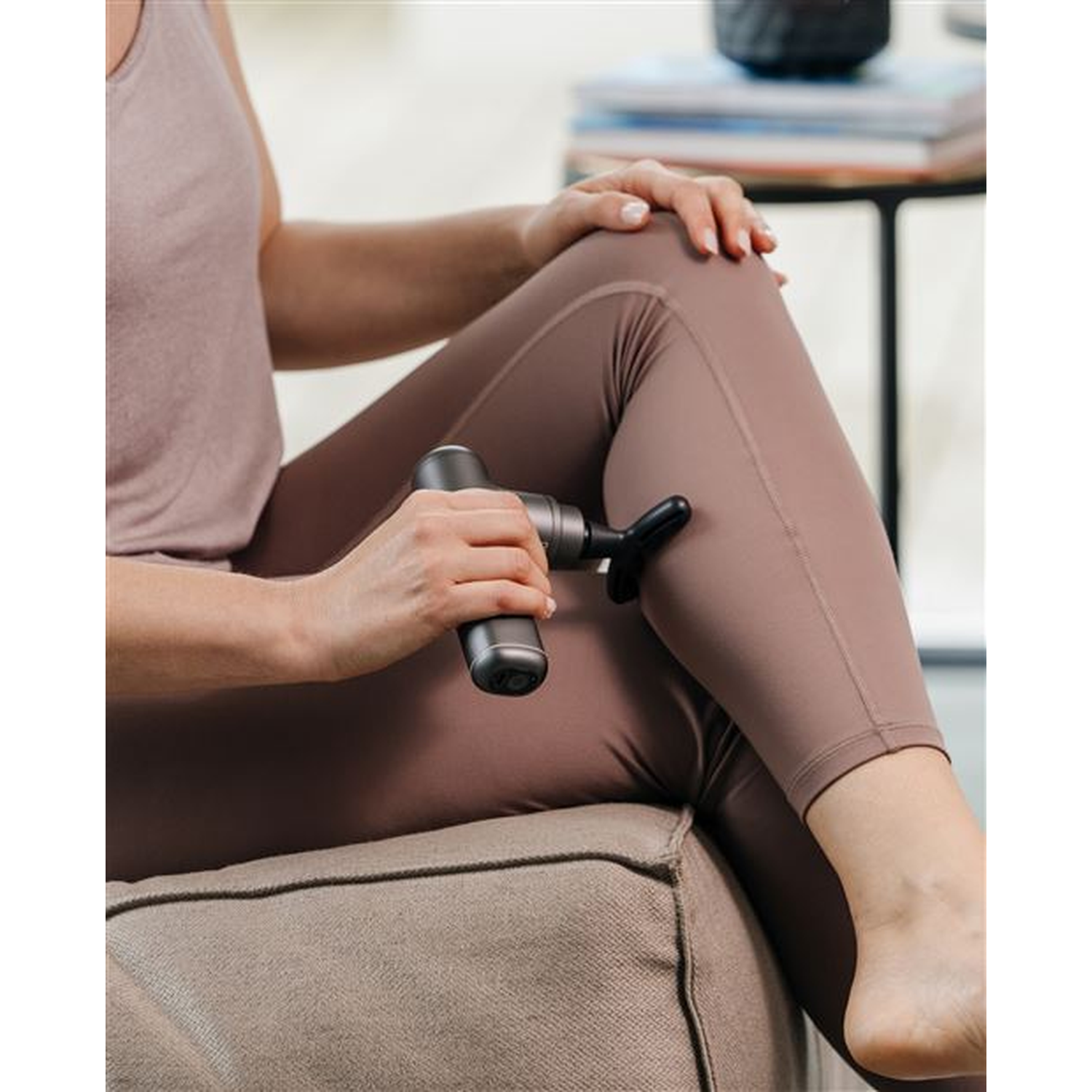 Une personne masse sa jambe avec un masseur manuel. Elle porte un legging et un débardeur, assise sur un canapé.