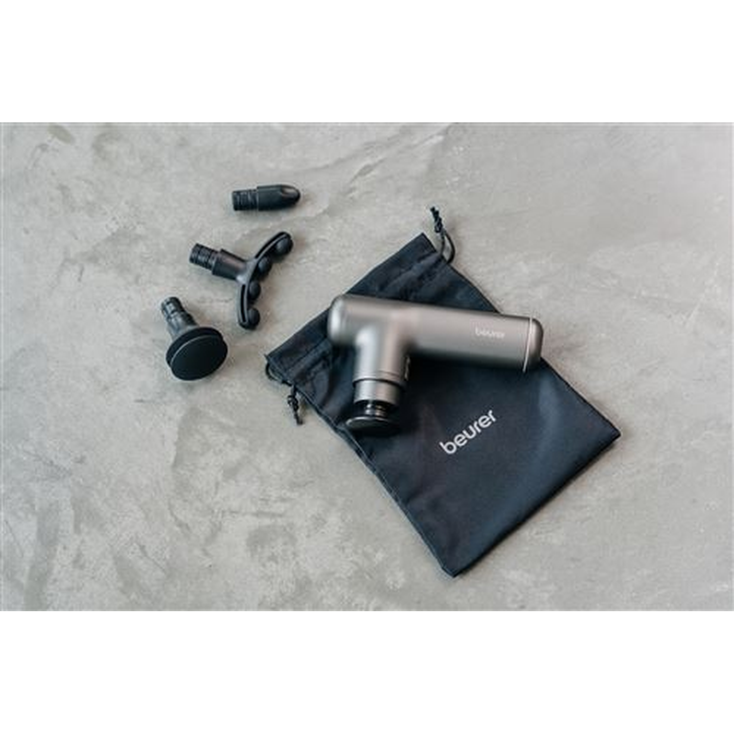 Un pistolet de massage gris avec accessoires sur une surface grise, à côté d'une pochette noire avec la marque 'beurer'.